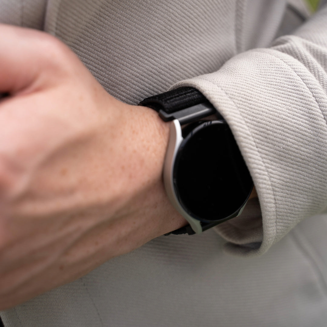 Das Handgelenk einer Person mit einer Smartwatch mit schwarzem Armband ist sichtbar. Die Person trägt eine hellfarbene Jacke. Das Zifferblatt der Uhr ist schwarz.