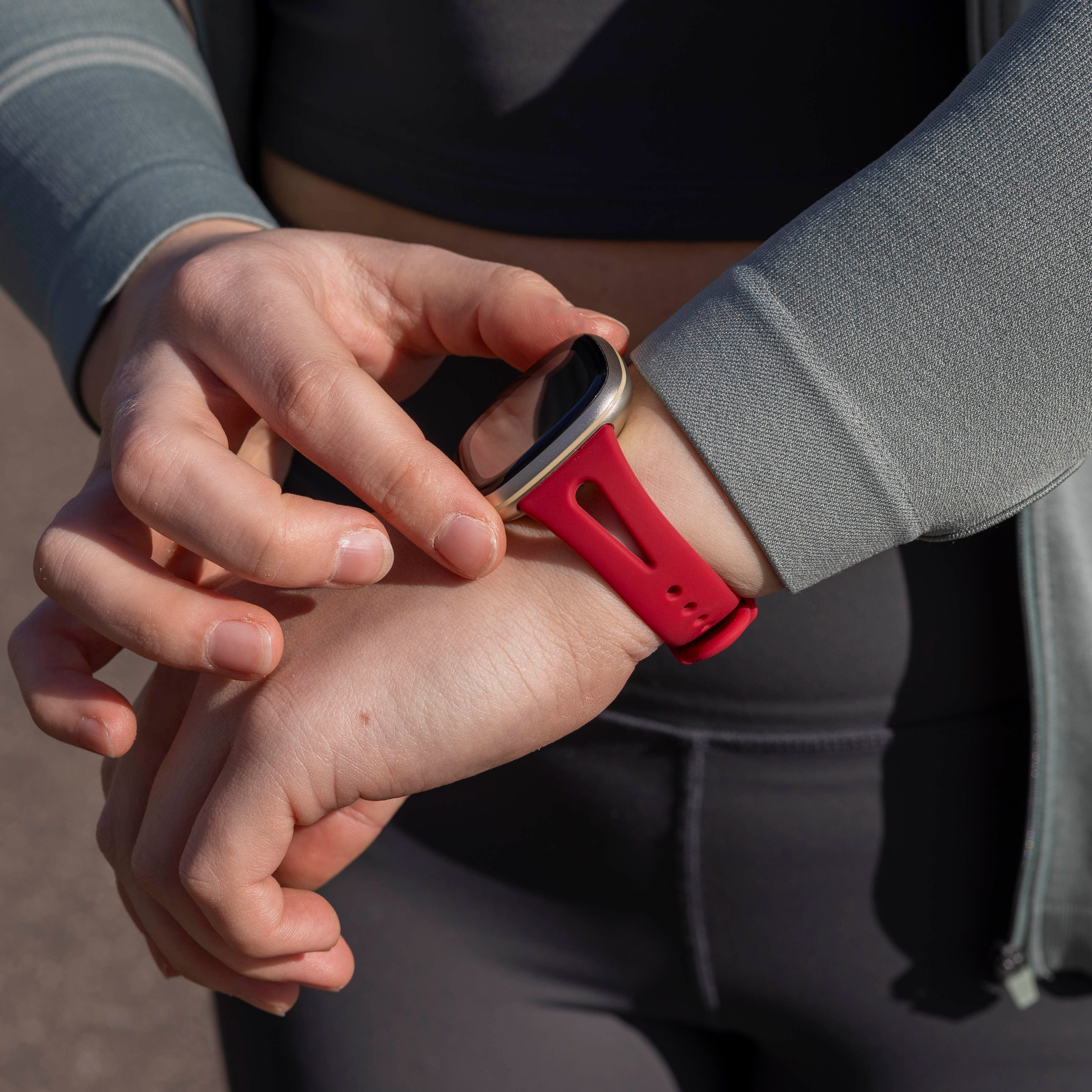 Die Hände einer Person, die eine Smartwatch mit rotem Armband einstellt. Sie trägt graue Sportkleidung und eine grüne Jacke. Der Hintergrund ist unscharf.