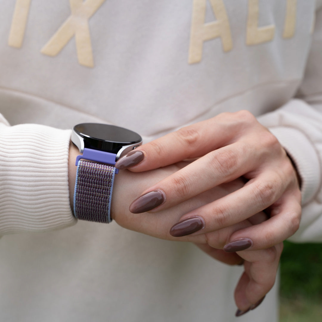 Eine Person, die eine Smartwatch mit lila Armband trägt. Die Person berührt die Uhr mit ihren braun lackierten Nägeln.