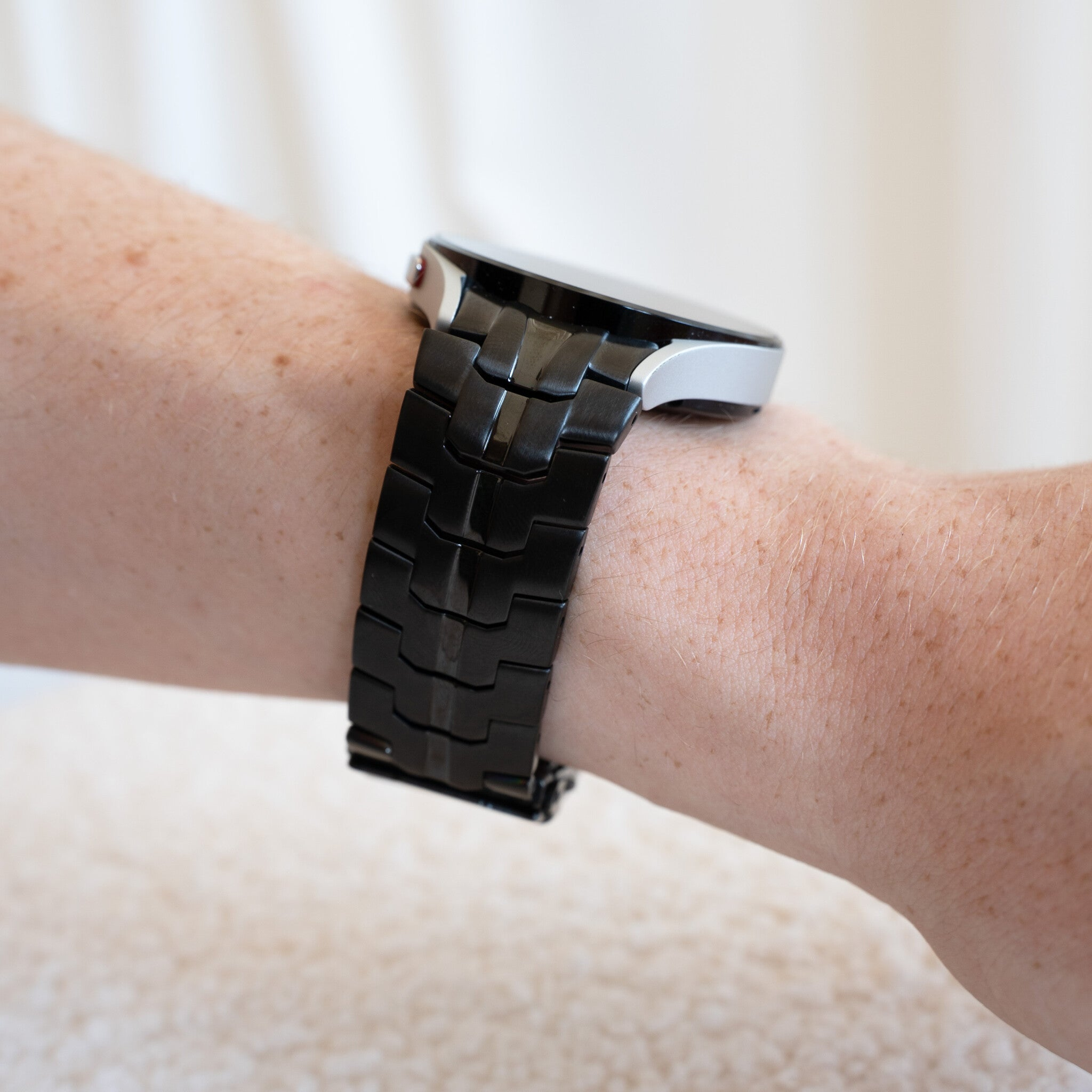 Ein Handgelenk mit einer schwarzen Uhr. Die Uhr hat ein schwarzes Armband und ein silbernes Zifferblatt. Die Haut hat Sommersprossen.