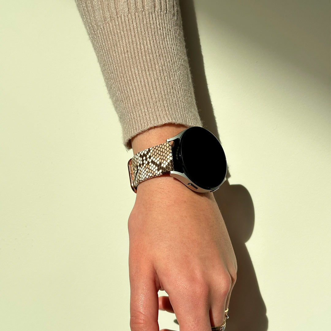 Handgelenk mit Smartwatch. Das Armband hat ein Schlangenmuster. Die Person trägt einen beigen Pullover.