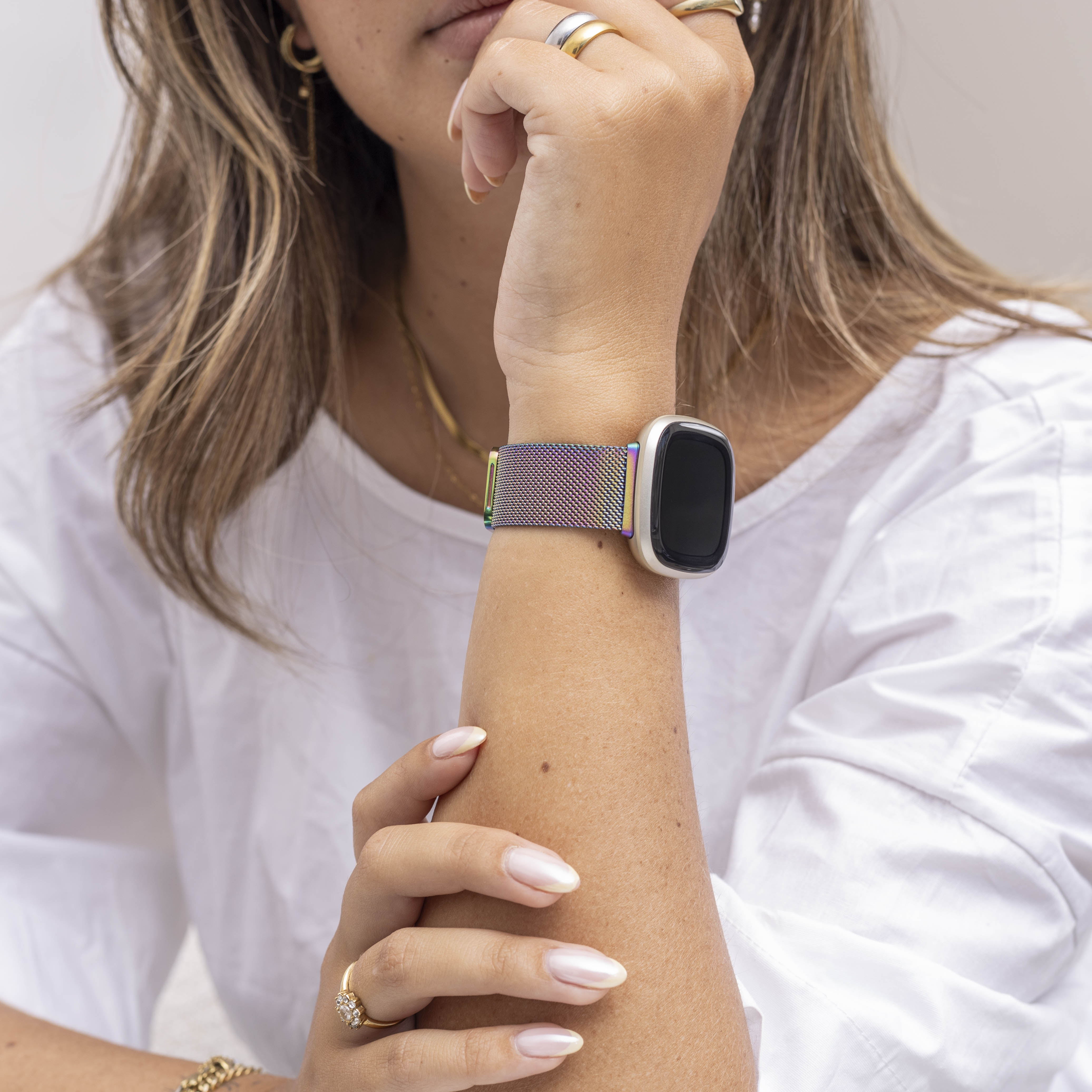 Das Handgelenk einer Frau mit einer Smartwatch mit einem regenbogenfarbenen Band. Sie trägt Ringe und ein weißes Hemd.