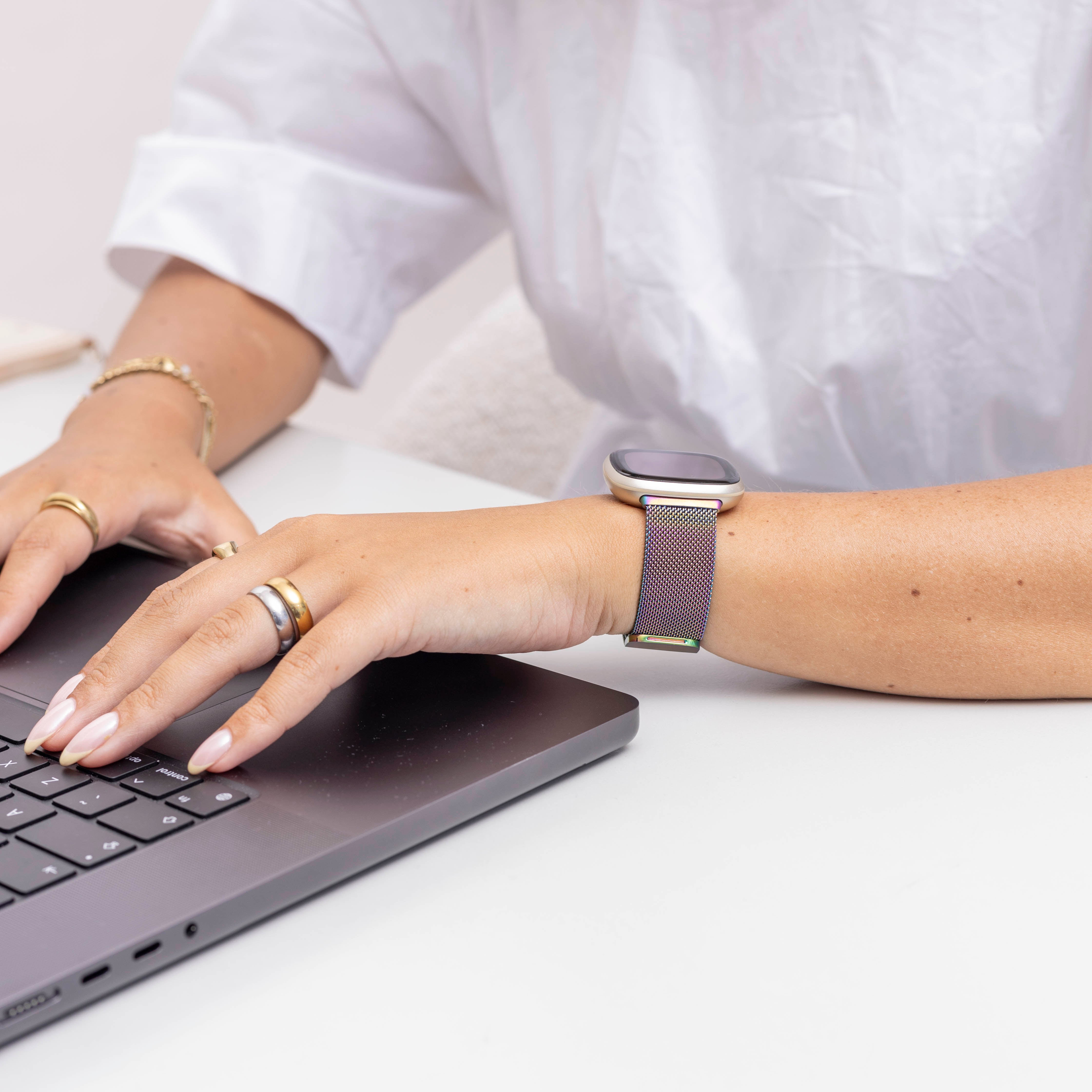 Hände einer Frau auf einem Laptop, mit Ringen und einer Uhr mit farbigem Armband, weißes Hemd.