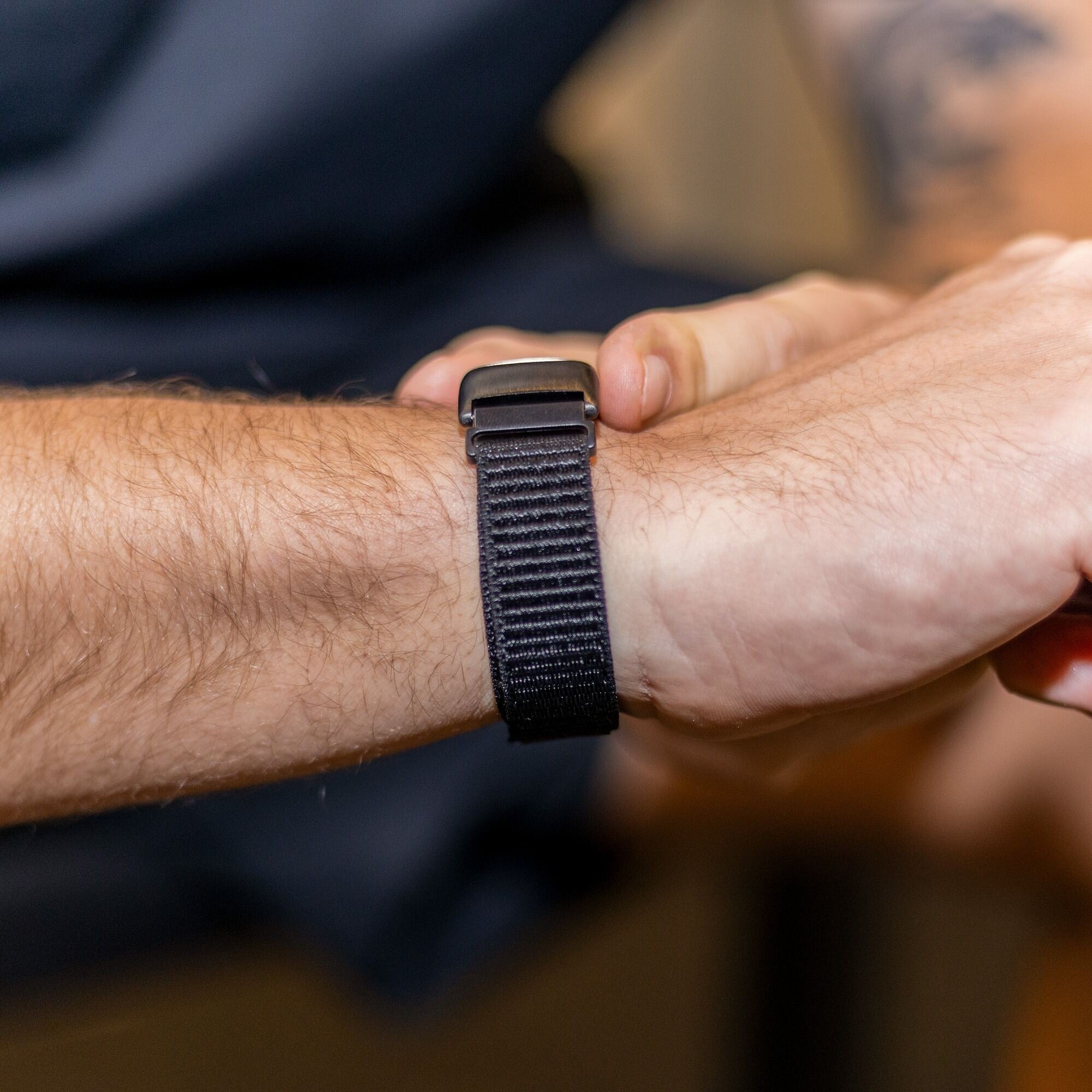 Nahaufnahme eines Handgelenks mit einem schwarzen Stoff-Uhrenarmband. Der Arm der Person hat helle Haut und etwas Haar.