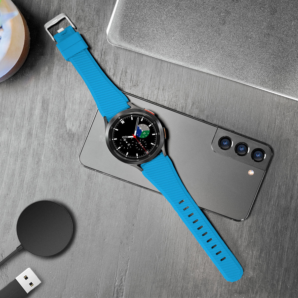 Eine schwarze Smartwatch mit blauem Armband liegt auf einem grauen Telefon. Ein Ladepad und ein Laptop sind in der Nähe, alles auf strukturierter Oberfläche.