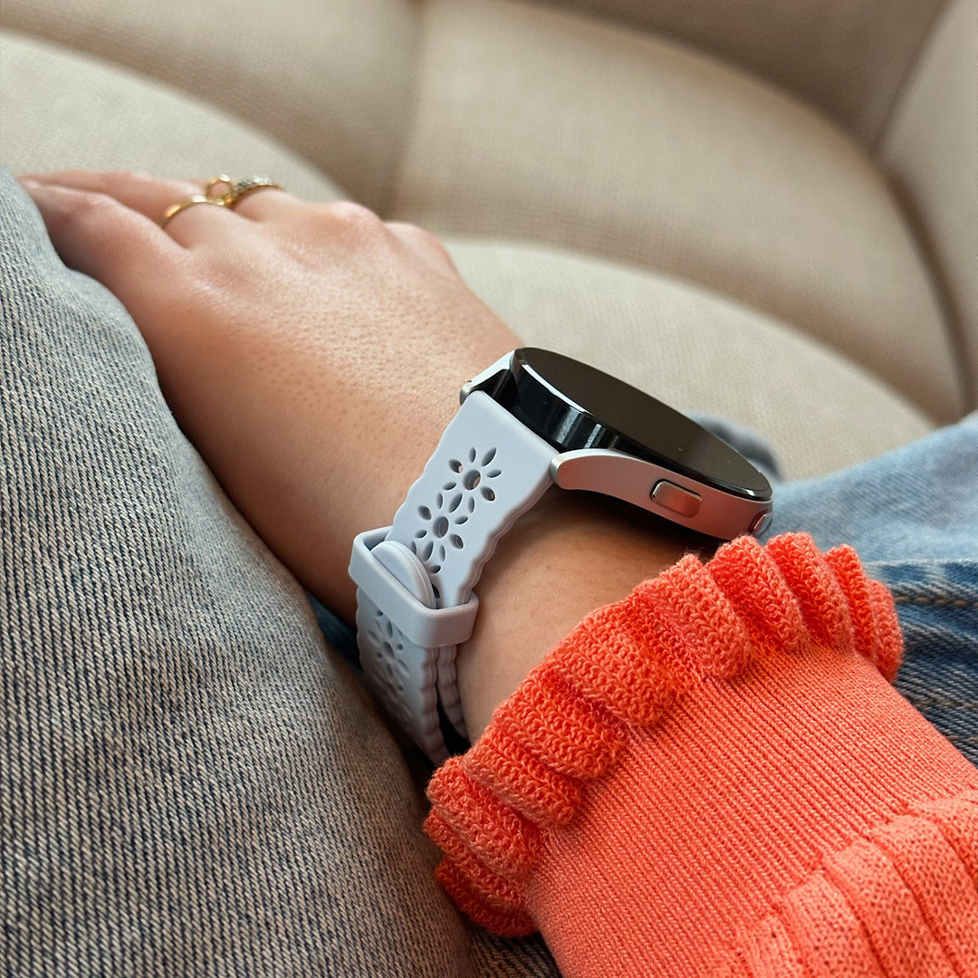Ein Arm mit Smartwatch und orangefarbenem Pullover. Die Uhr hat ein hellblaues Armband. Die Person trägt Ringe.