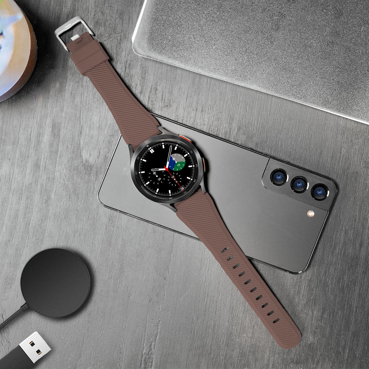 Eine braune Smartwatch mit schwarzem Zifferblatt und braunem Armband liegt auf einem Telefon. Ein Ladegerät, ein Laptop und ein Untersetzer sind ebenfalls sichtbar.
