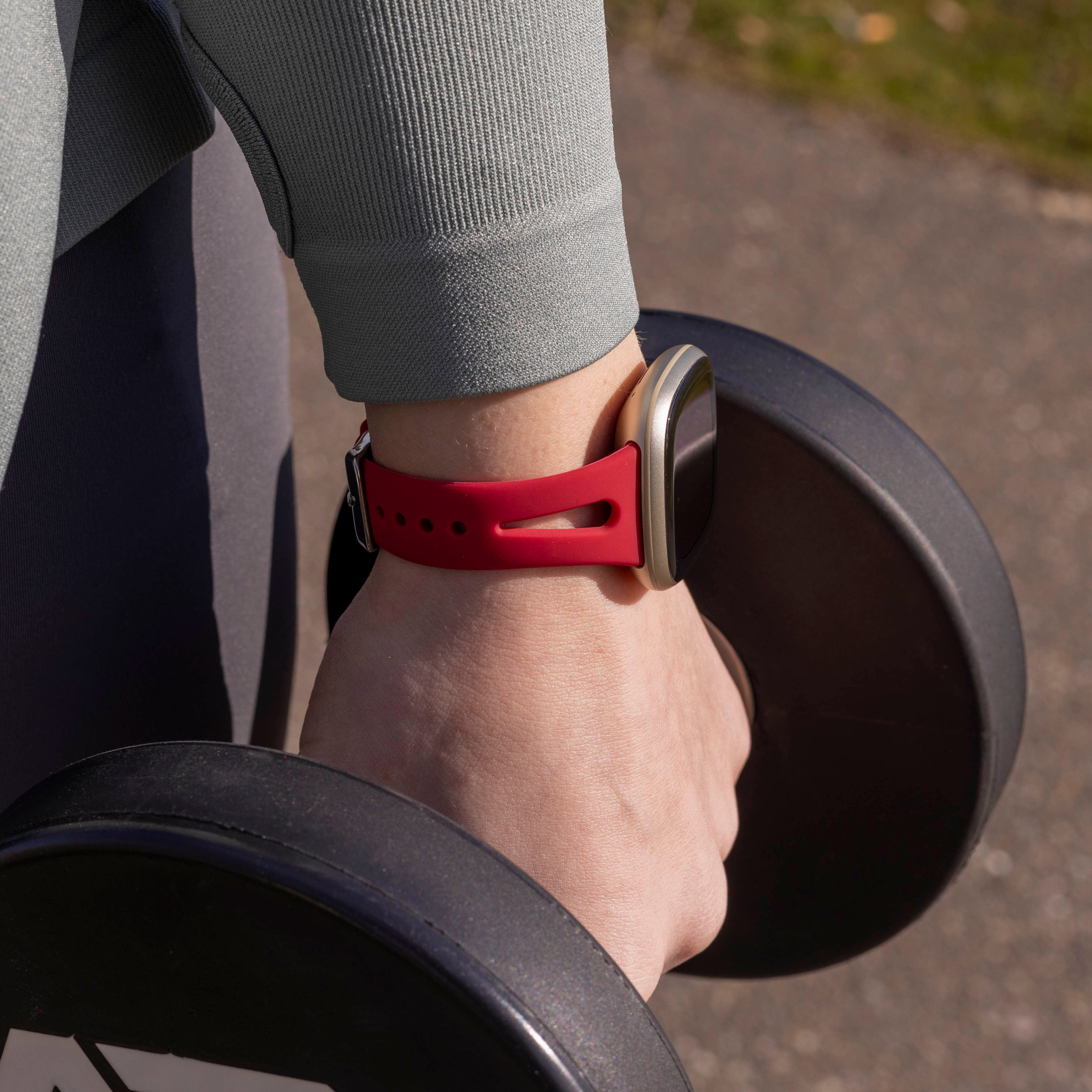 Der Arm einer Person hält eine Hantel, an der eine rote Uhr getragen wird. Die Person trägt einen grauen Pullover. Der Hintergrund ist unscharf.
