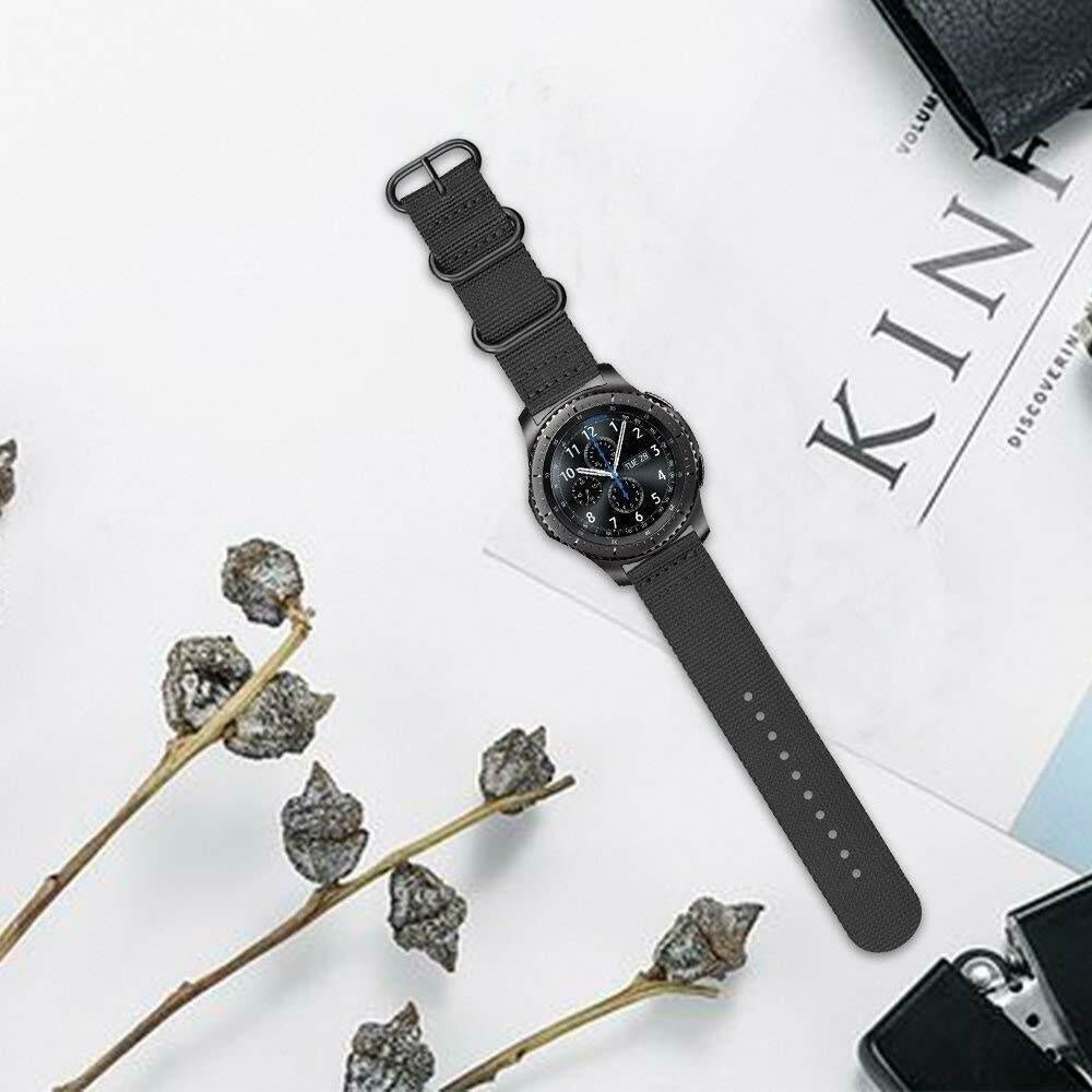 Eine schwarze Smartwatch mit schwarzem Armband liegt auf einer weißen Oberfläche. Getrocknete Pflanzen und eine Zeitschrift sind ebenfalls zu sehen.