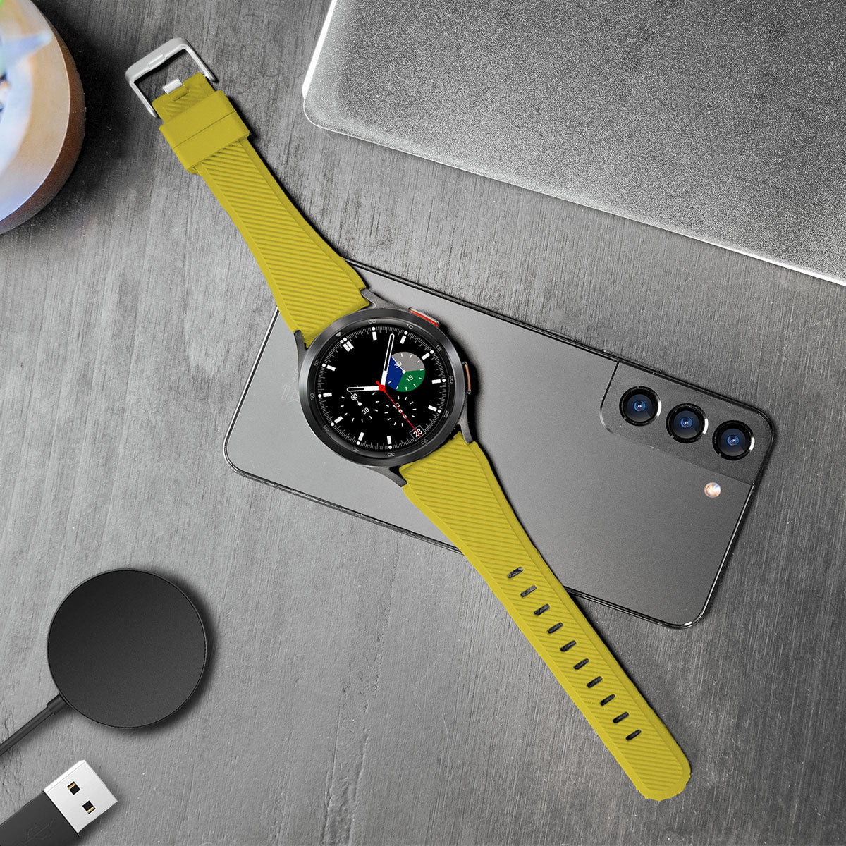 Eine gelbe Smartwatch liegt auf einem Telefon. Ein kabelloses Ladegerät und ein Laptop befinden sich in der Nähe auf einer grauen Oberfläche.