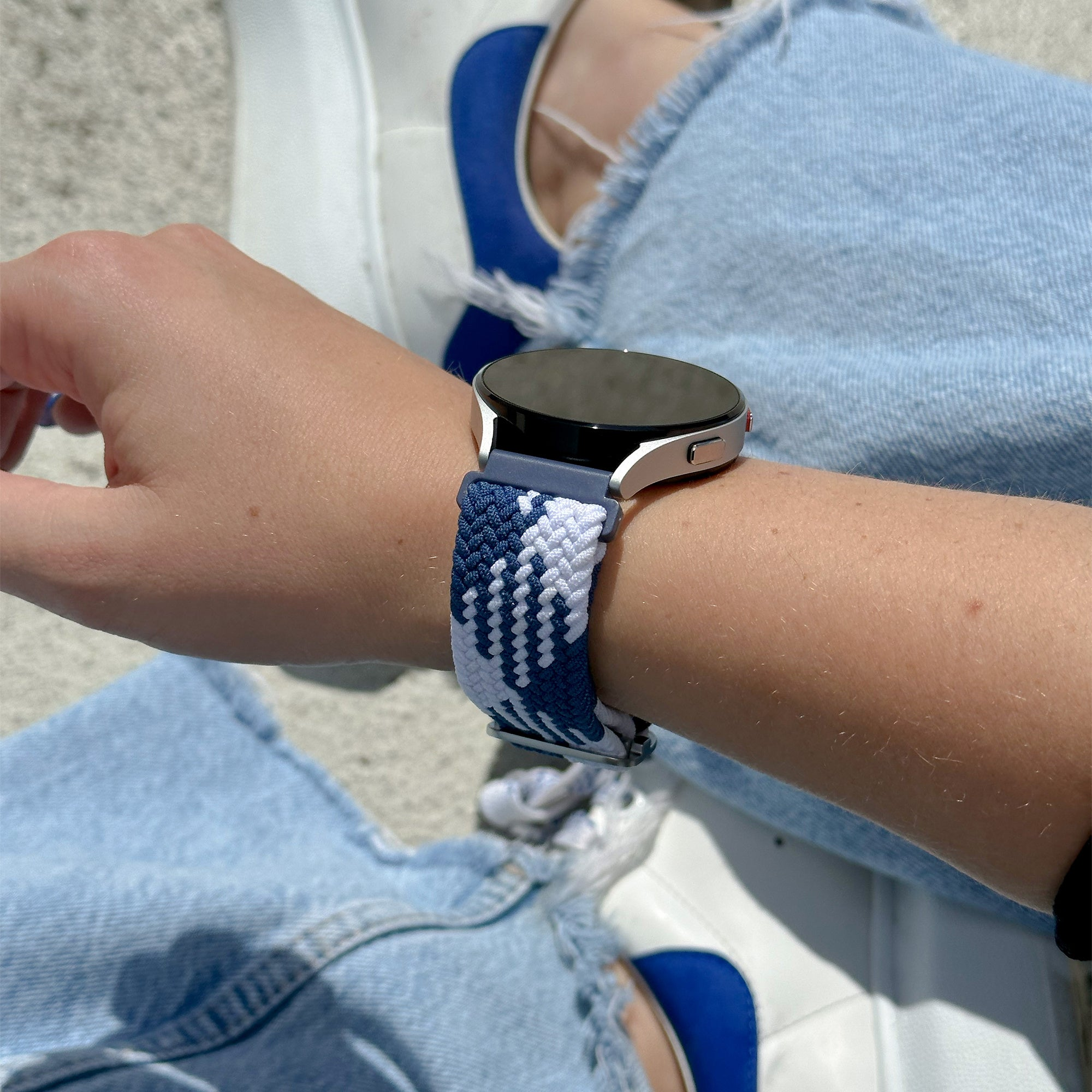 Handgelenk mit Smartwatch. Das Armband ist blau-weiß geflochten. Die Person trägt Jeans und weiße Schuhe.