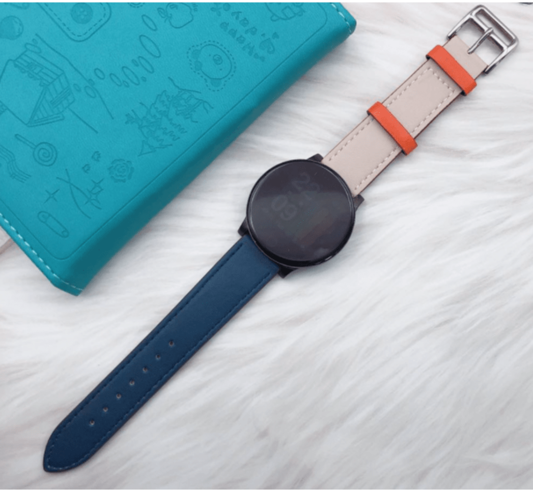Eine schwarze Smartwatch mit blauem und beigem Armband liegt auf einer weißen Pelzoberfläche neben einem türkisfarbenen Notizbuch.