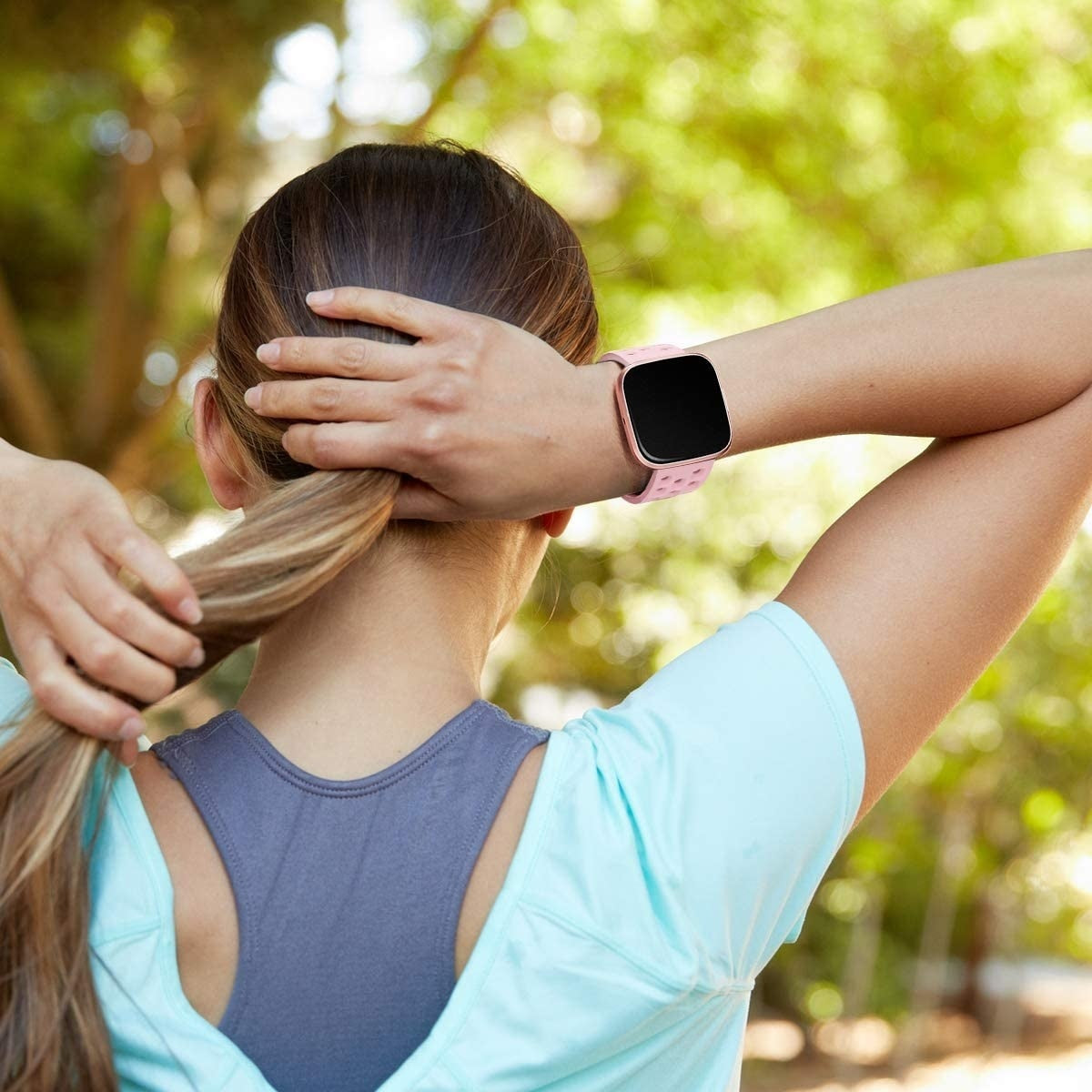 Frau von hinten mit Smartwatch, die ihren Pferdeschwanz richtet. Sie trägt ein hellblaues Shirt und grauen Sport-BH, mit grünen Bäumen im Hintergrund.