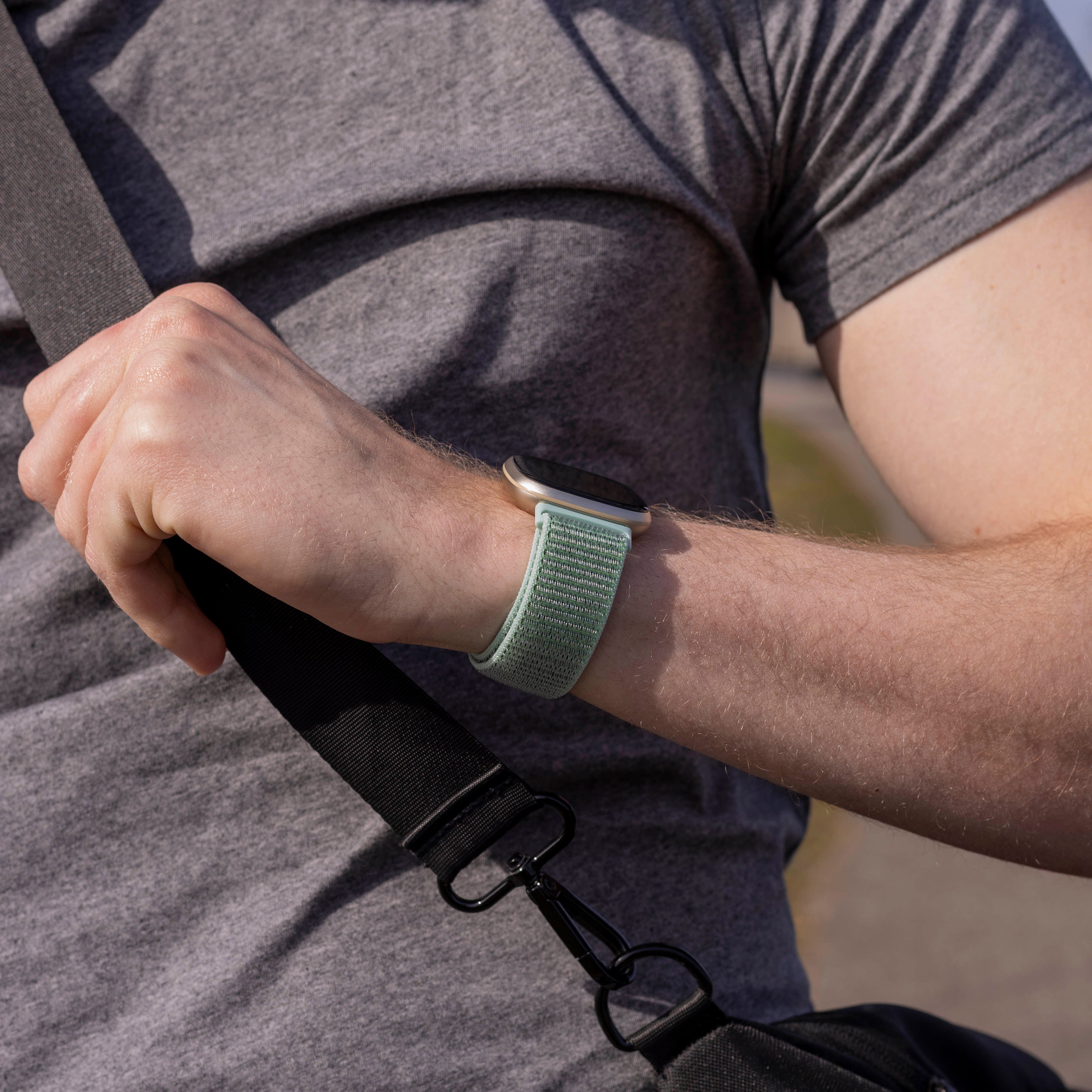 Ein Arm mit Uhr und grünem Band, hält einen schwarzen Taschenriemen. Die Person trägt ein graues Hemd.
