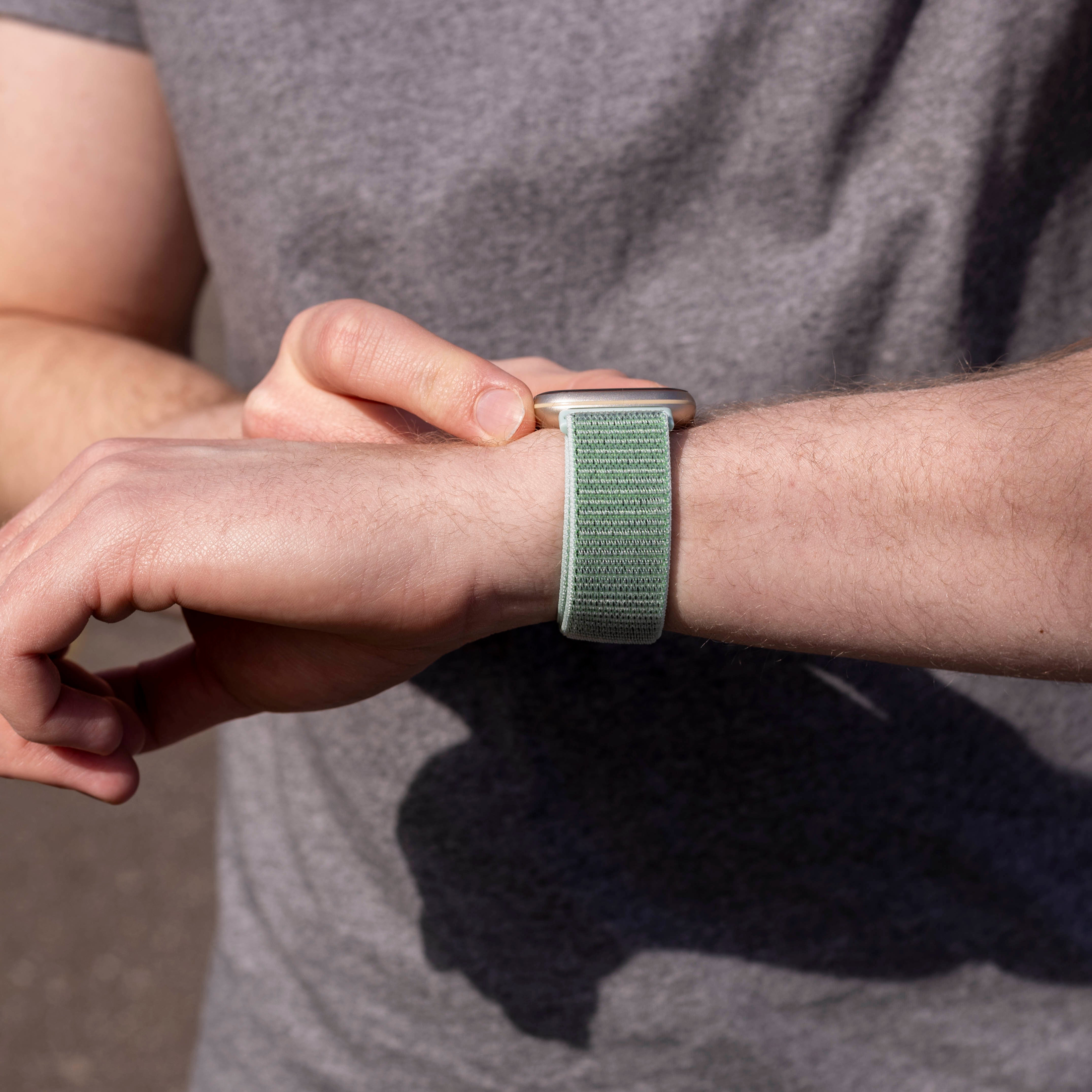 Eine Person trägt ein graues T-Shirt und überprüft eine grüne Uhr an ihrem Handgelenk. Das Armband ist hellgrün.