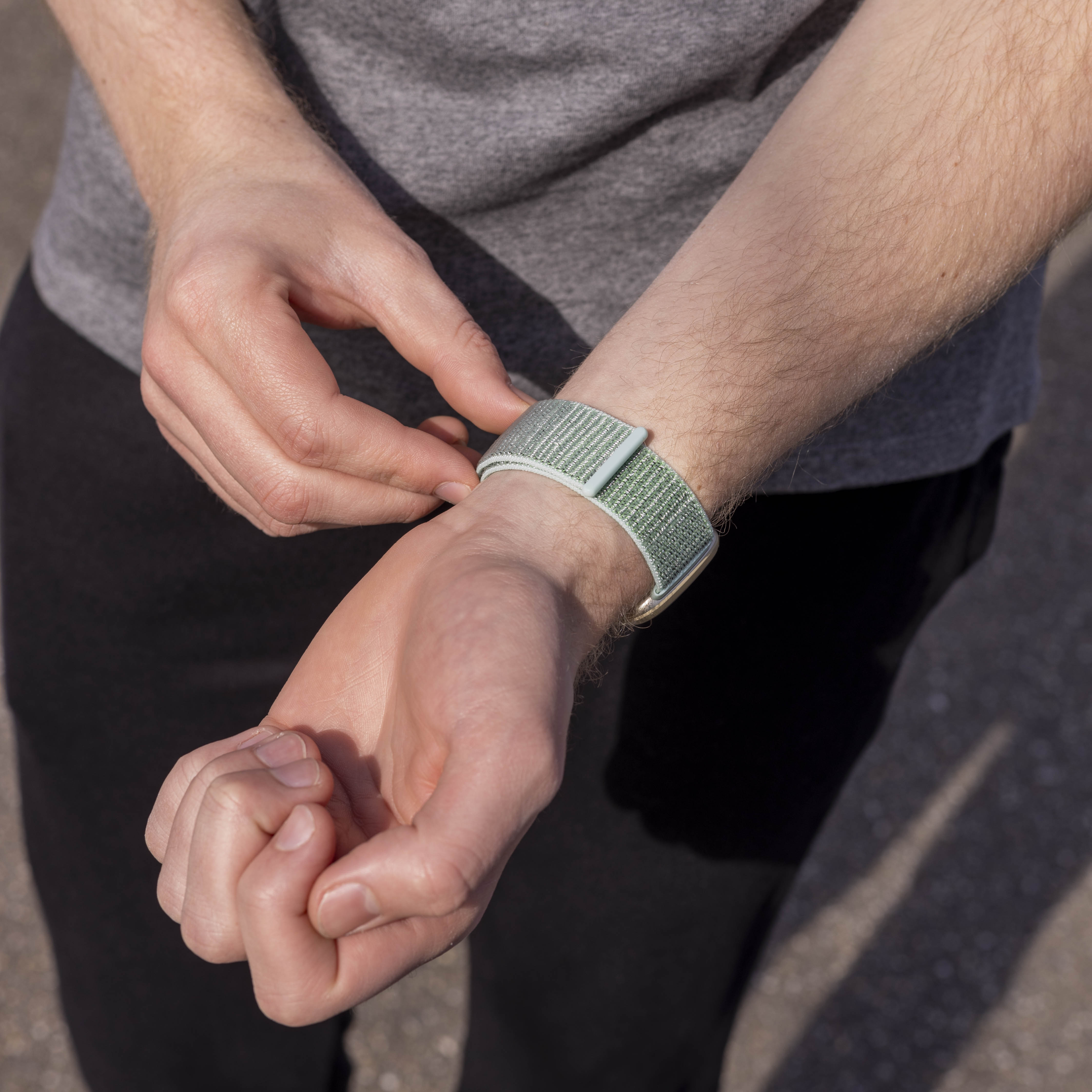 Eine Person, die ein hellgrünes Armband an ihrem Handgelenk anpasst. Sie trägt ein graues Hemd und eine schwarze Hose, draußen.