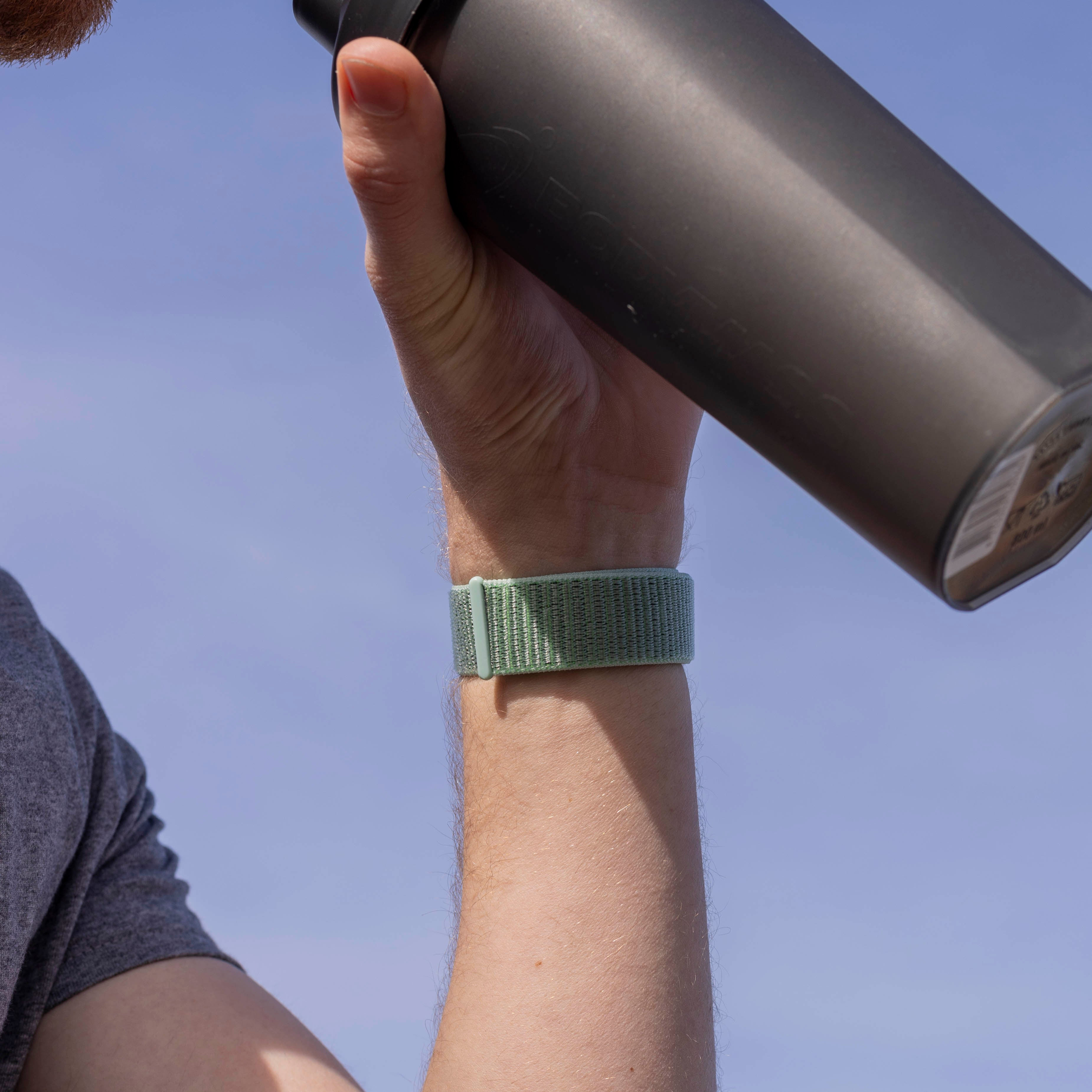 Der Arm einer Person mit grünem Armband hält eine schwarze Wasserflasche vor blauem Himmel.