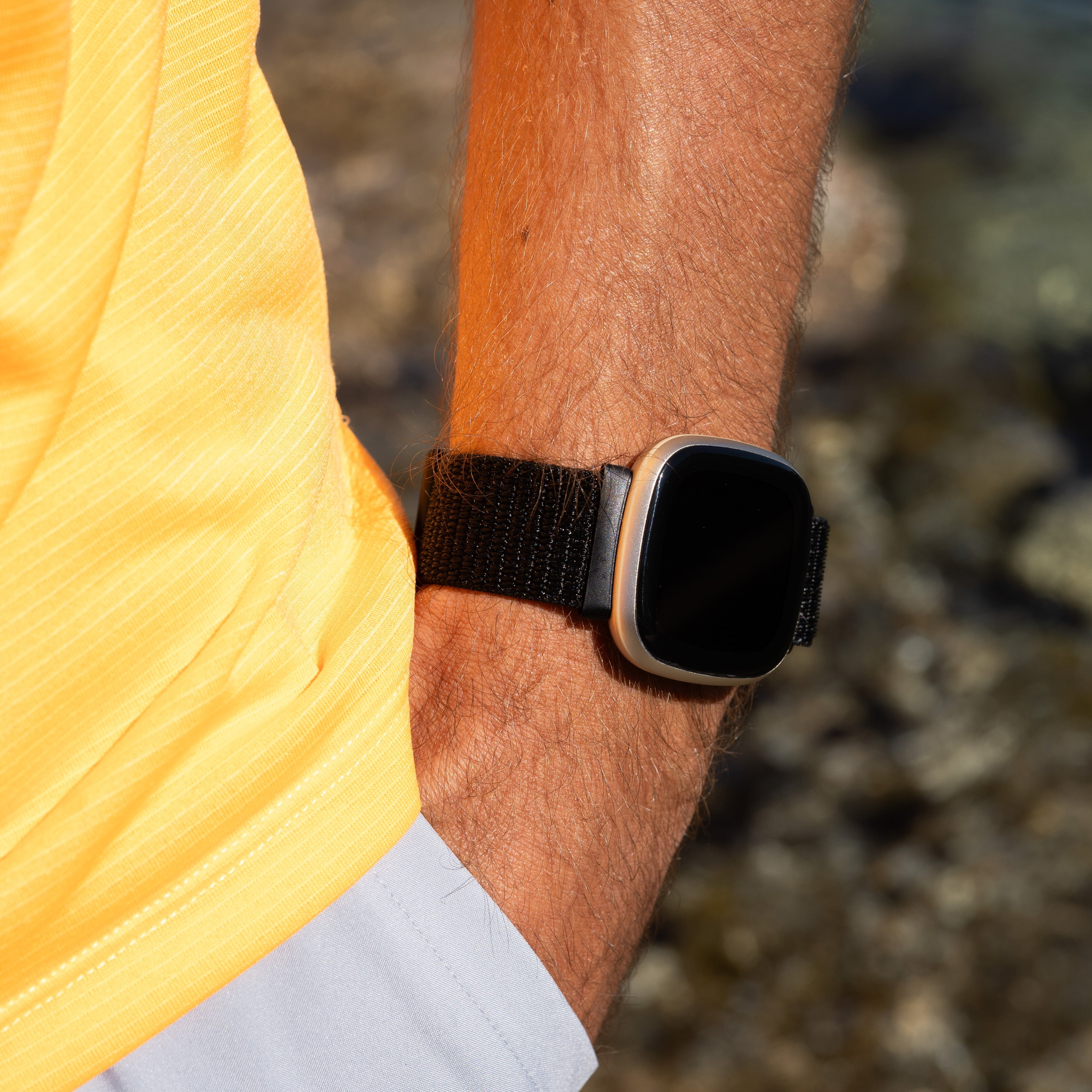Nahaufnahme eines Mannes mit Smartwatch. Er trägt ein gelbes Hemd. Der Hintergrund zeigt Wasser und Felsen.