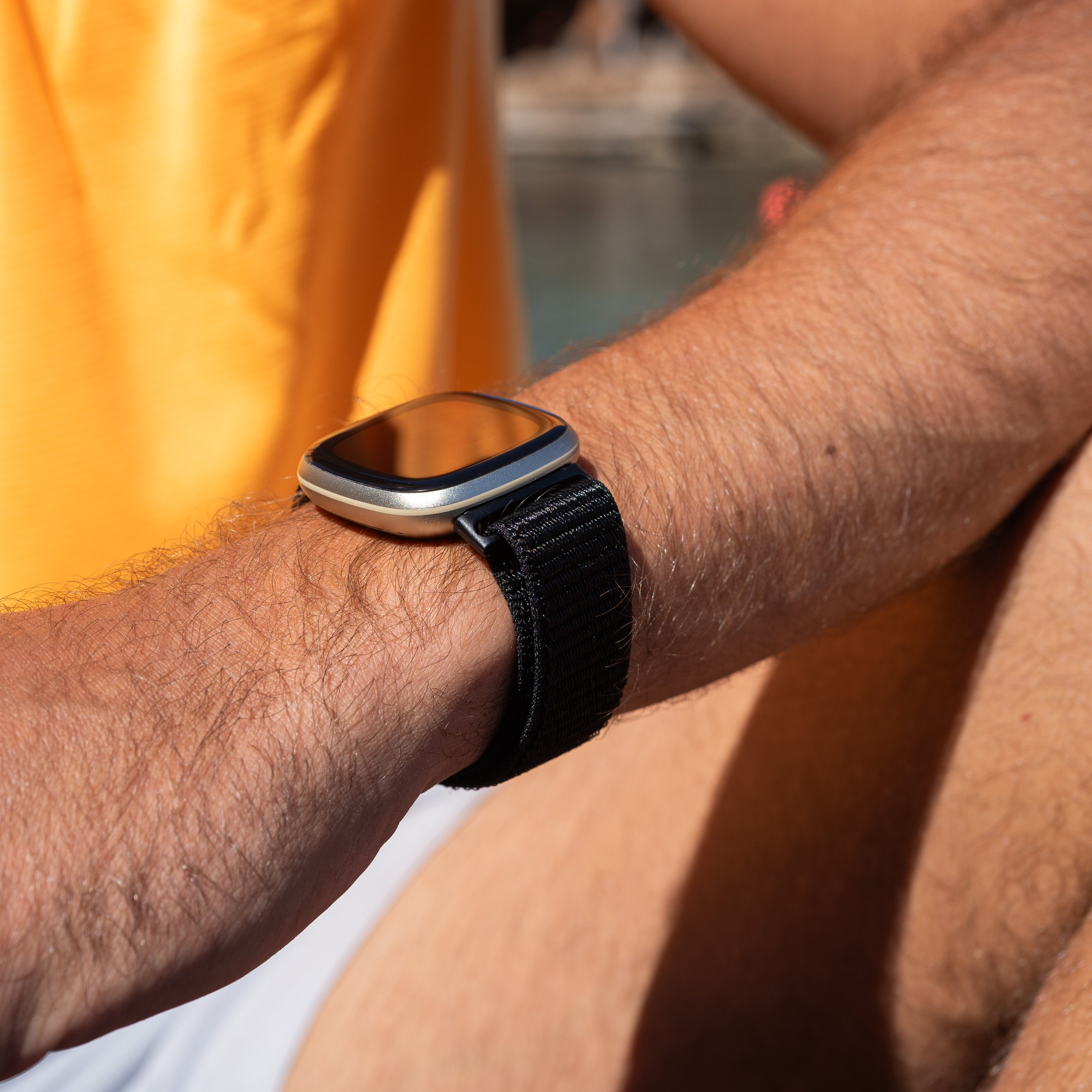 Arm einer Person mit einer Uhr. Die Uhr hat ein schwarzes Armband und ein silbernes Gehäuse. Die Person trägt ein orangefarbenes Hemd.
