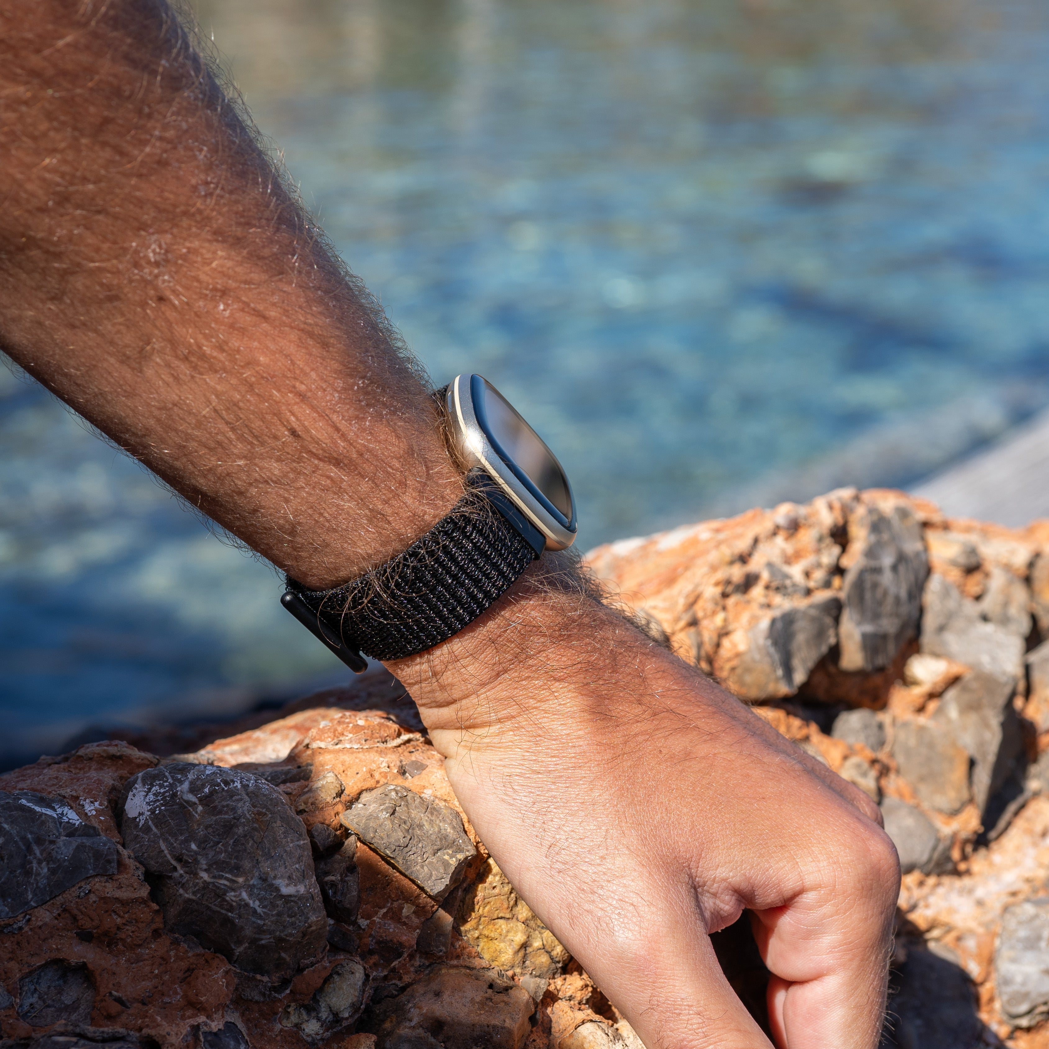 Ein Handgelenk mit einer Uhr ruht auf Felsen. Der Hintergrund zeigt verschwommenes Wasser. Die Uhr hat ein schwarzes Armband.