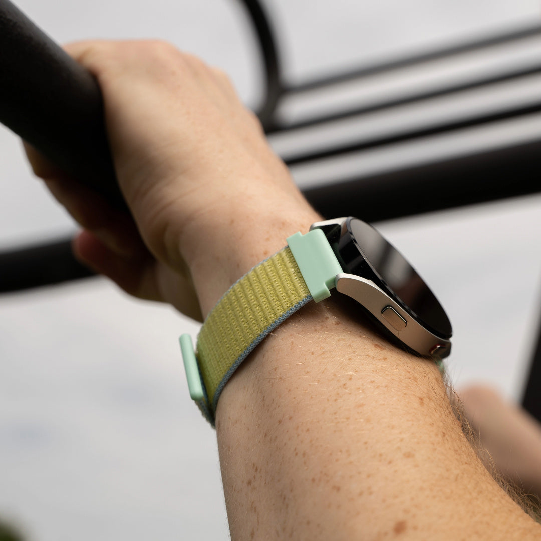 Handgelenk mit Smartwatch. Die Uhr hat ein gelbes Armband mit grünem Verschluss. Die Person hält eine schwarze Stange.