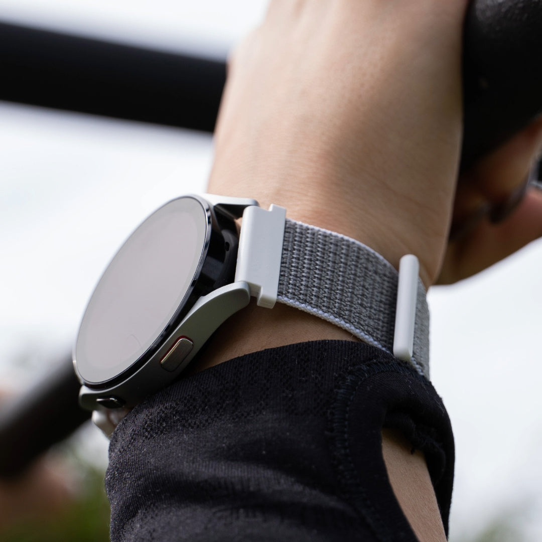 Das Handgelenk einer Person mit einer silbernen Smartwatch und einem grauen Armband. Die Person trägt einen schwarzen Ärmel. Der Hintergrund ist unscharf.