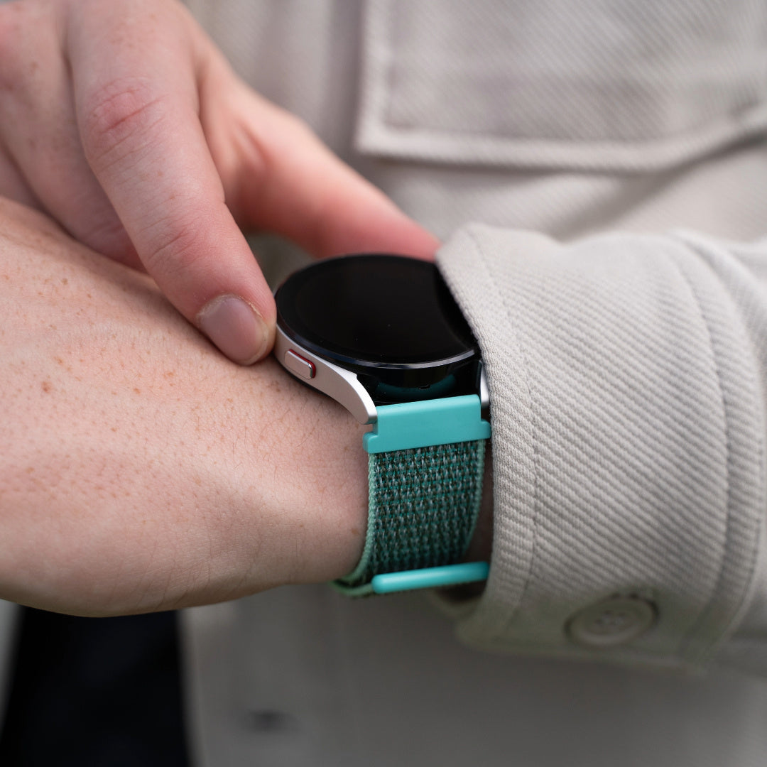 Eine Person trägt eine Smartwatch mit grünem Armband. Die Hand der Person stellt die Uhr ein. Beigefarbene Jacke im Hintergrund.