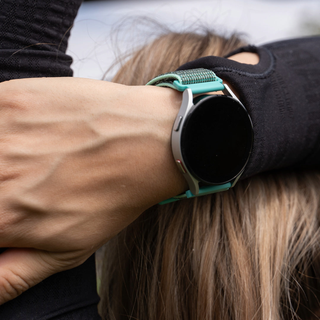 Eine Person trägt eine silberne Smartwatch mit schwarzem Bildschirm und türkisfarbenem Armband, auf langen braunen Haaren, in einem schwarzen Hemd.