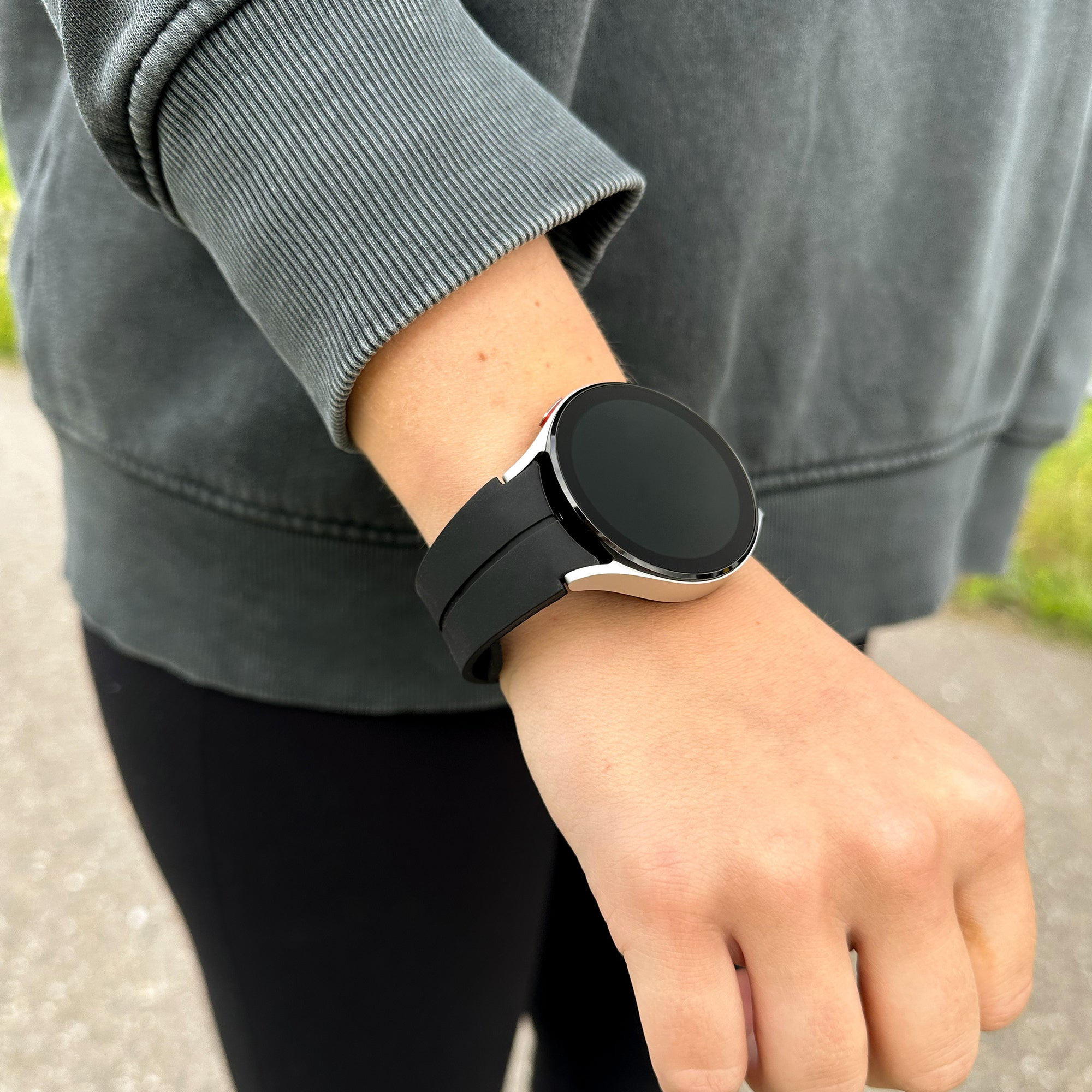 Das Handgelenk einer Person mit einer schwarzen Smartwatch. Die Person trägt einen grauen Pullover und schwarze Leggings. Hintergrund im Freien.