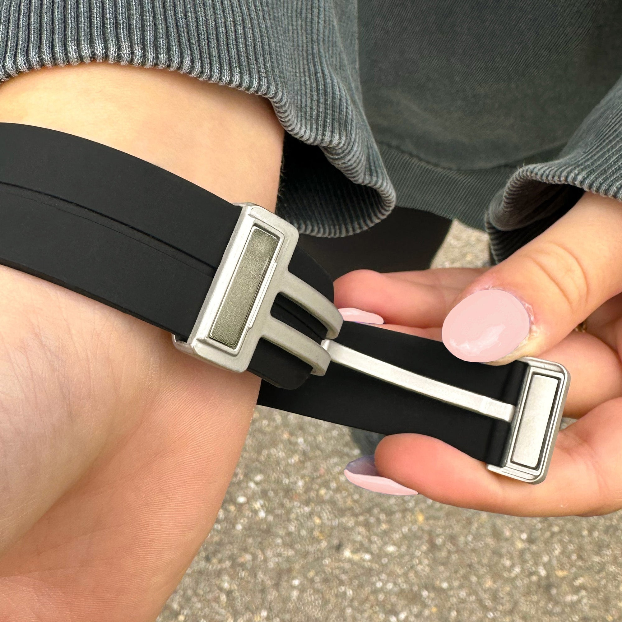 Das Handgelenk einer Person mit einem schwarzen Armband. Die Hände der Person halten das Armband. Die Nägel sind pink lackiert.