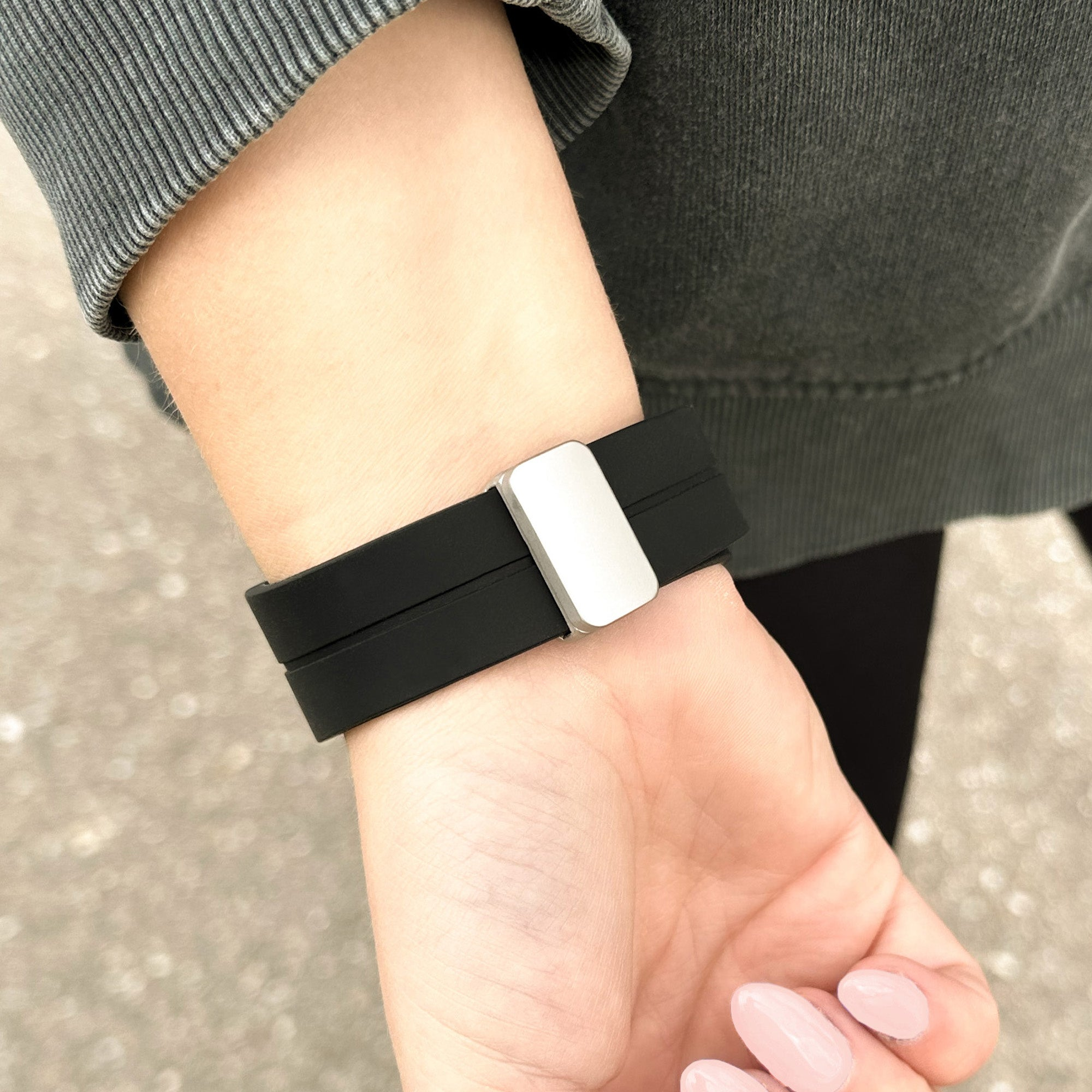 Handgelenk mit schwarzer Armbanduhr, silberner Schließe und rosafarbenem Nagellack. Graue Kleidung sichtbar.