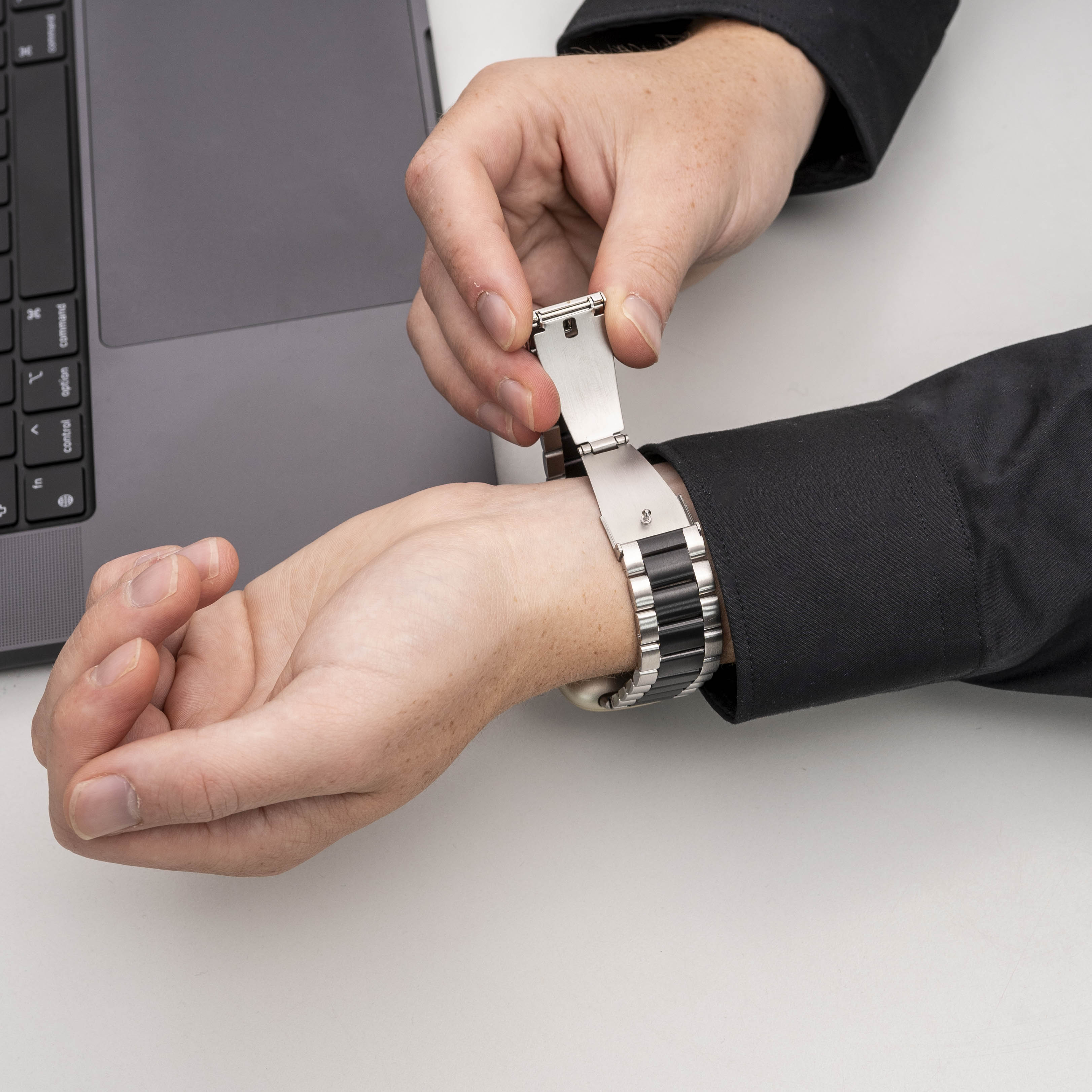 Hände einer Person, die eine Armbanduhr einstellt. Im Hintergrund ein Laptop. Die Person trägt ein schwarzes Hemd.