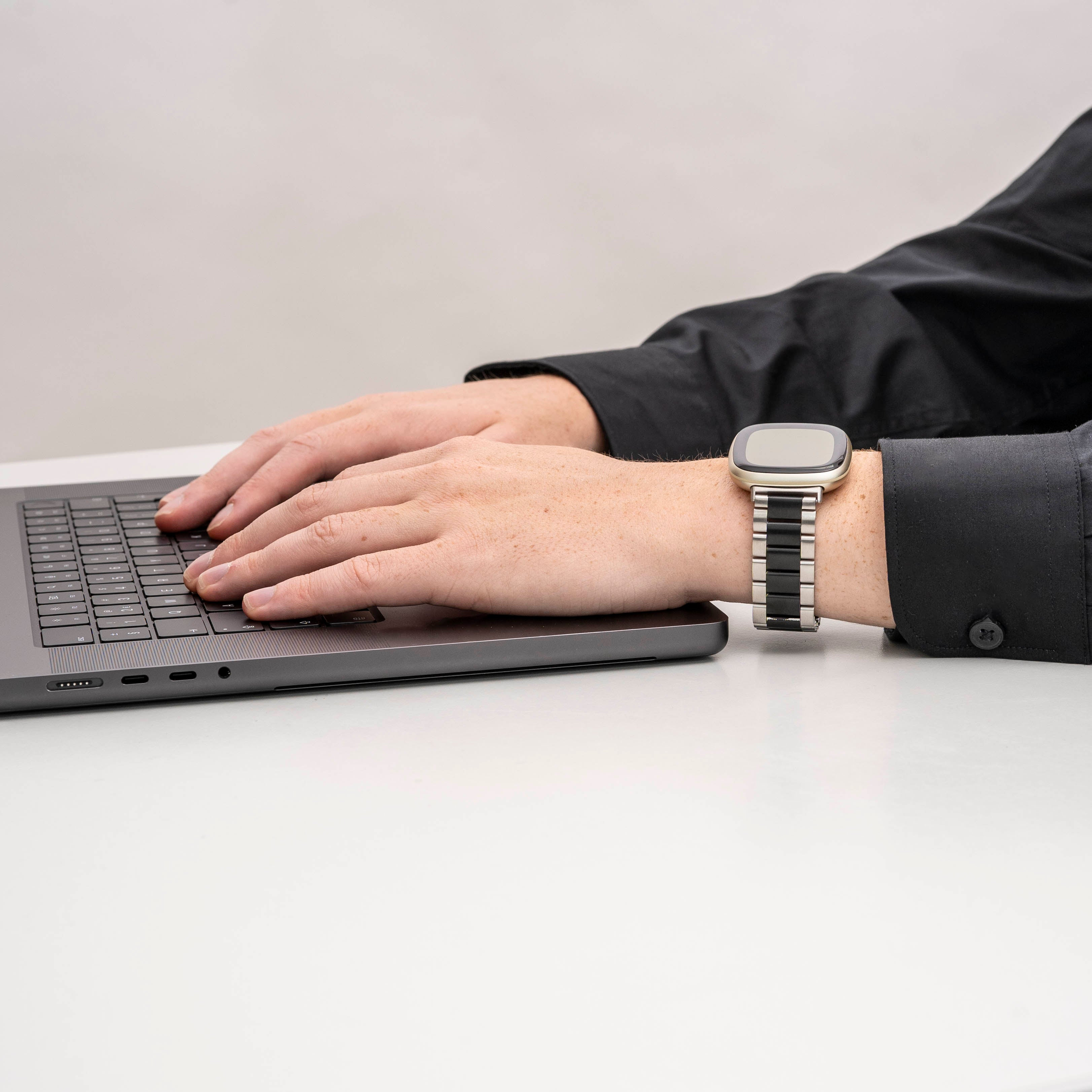 Hände tippen auf einem Laptop. Eine Uhr befindet sich am linken Handgelenk. Schwarzes Hemd. Weißer Tisch und Hintergrund.