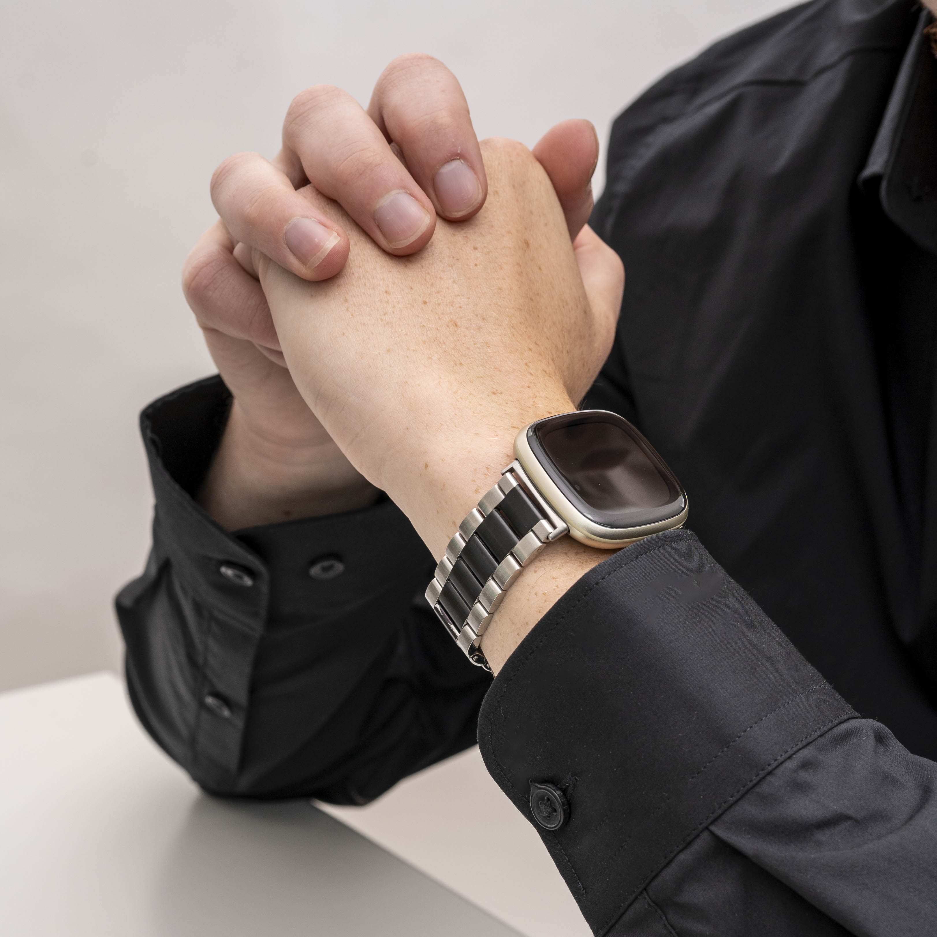 Hände gefaltet, Armbanduhr mit schwarz-silbernem Band, schwarzes Hemd. Hintergrund ist eine weiße Oberfläche.
