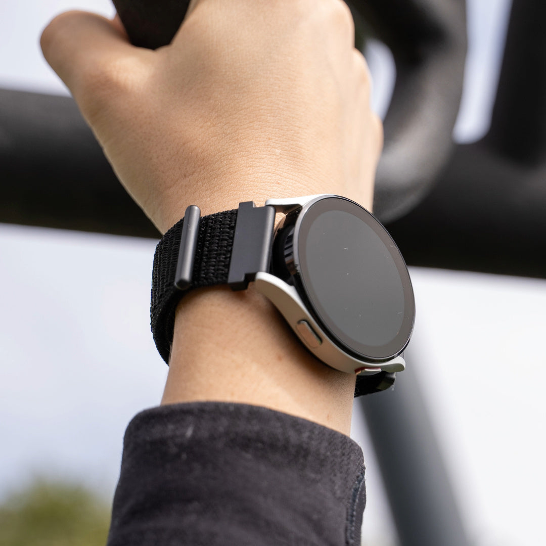 Das Handgelenk einer Person mit einer silbernen Smartwatch mit schwarzem Armband. Die Person trägt ein schwarzes Langarmshirt. Der Hintergrund ist unscharf.