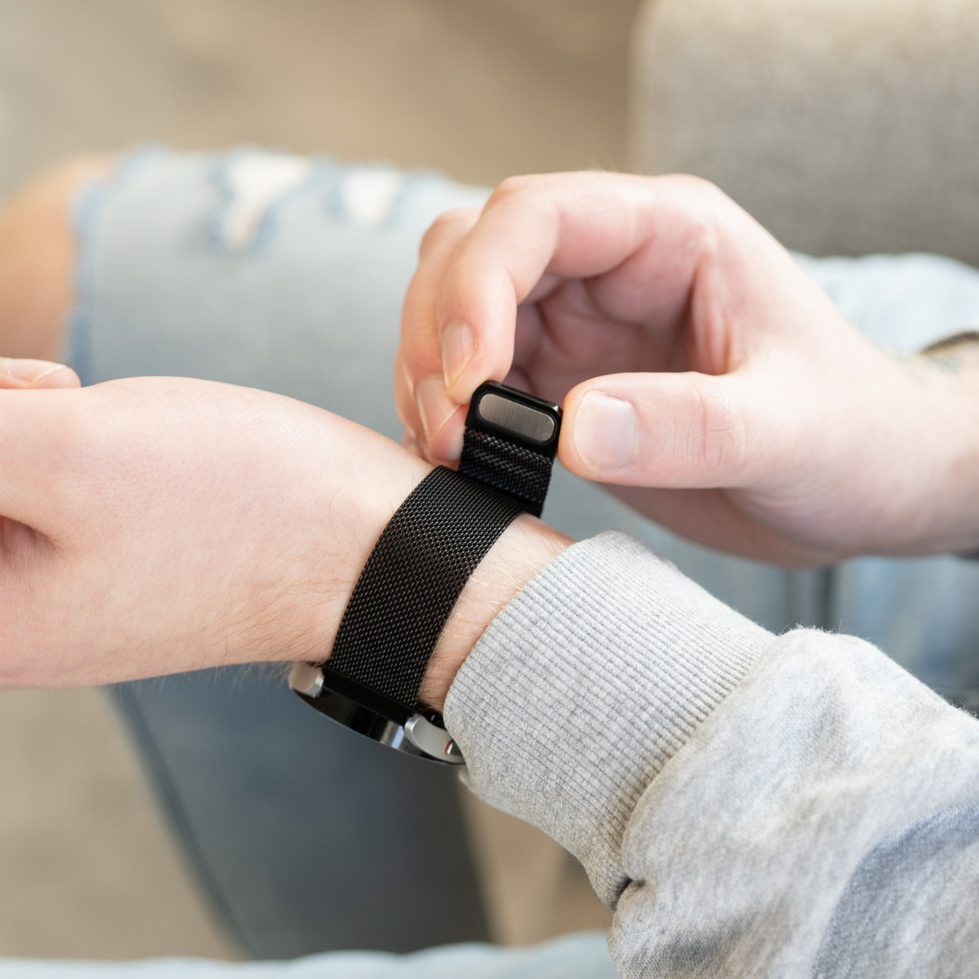 Eine Person befestigt ein schwarzes Uhrenarmband an ihrem Handgelenk. Die Uhr hat ein schwarzes Mesh-Armband, und die Person trägt einen grauen Pullover.
