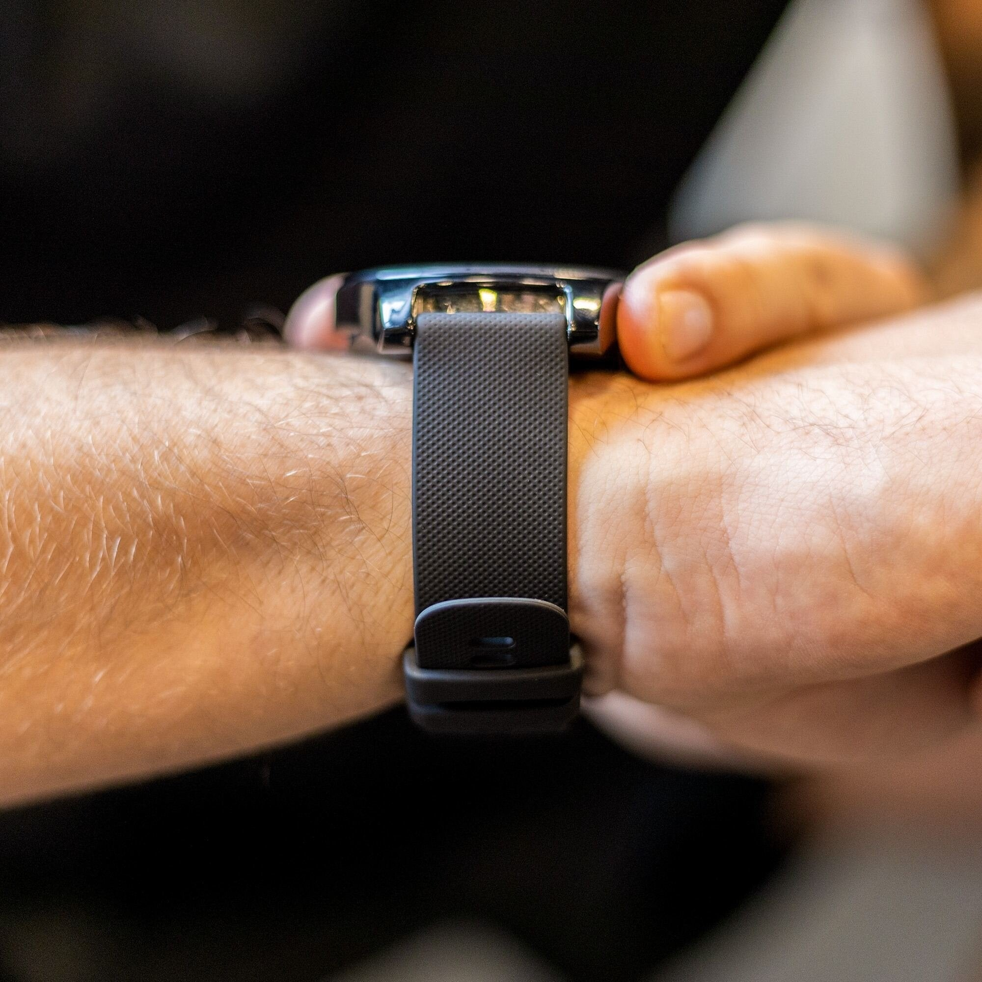 Nahaufnahme eines Handgelenks mit dunkler grauer Smartwatch und Armband. Haut ist sichtbar. Der Hintergrund ist verschwommen und dunkel.