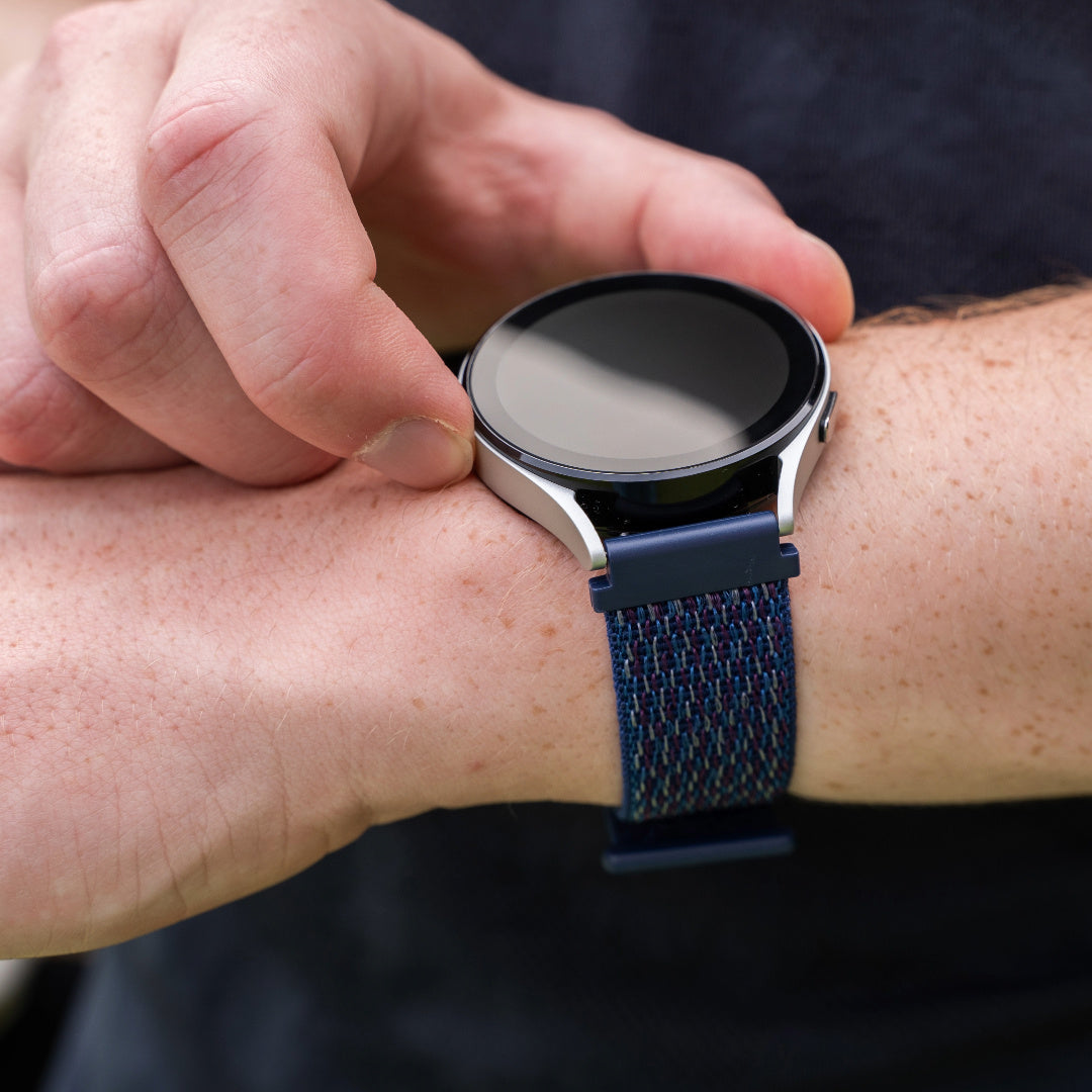 Eine Person trägt eine Smartwatch. Die Uhr hat ein silbernes Gehäuse und ein blaues Stoffarmband. Die Hand der Person stellt die Uhr ein.