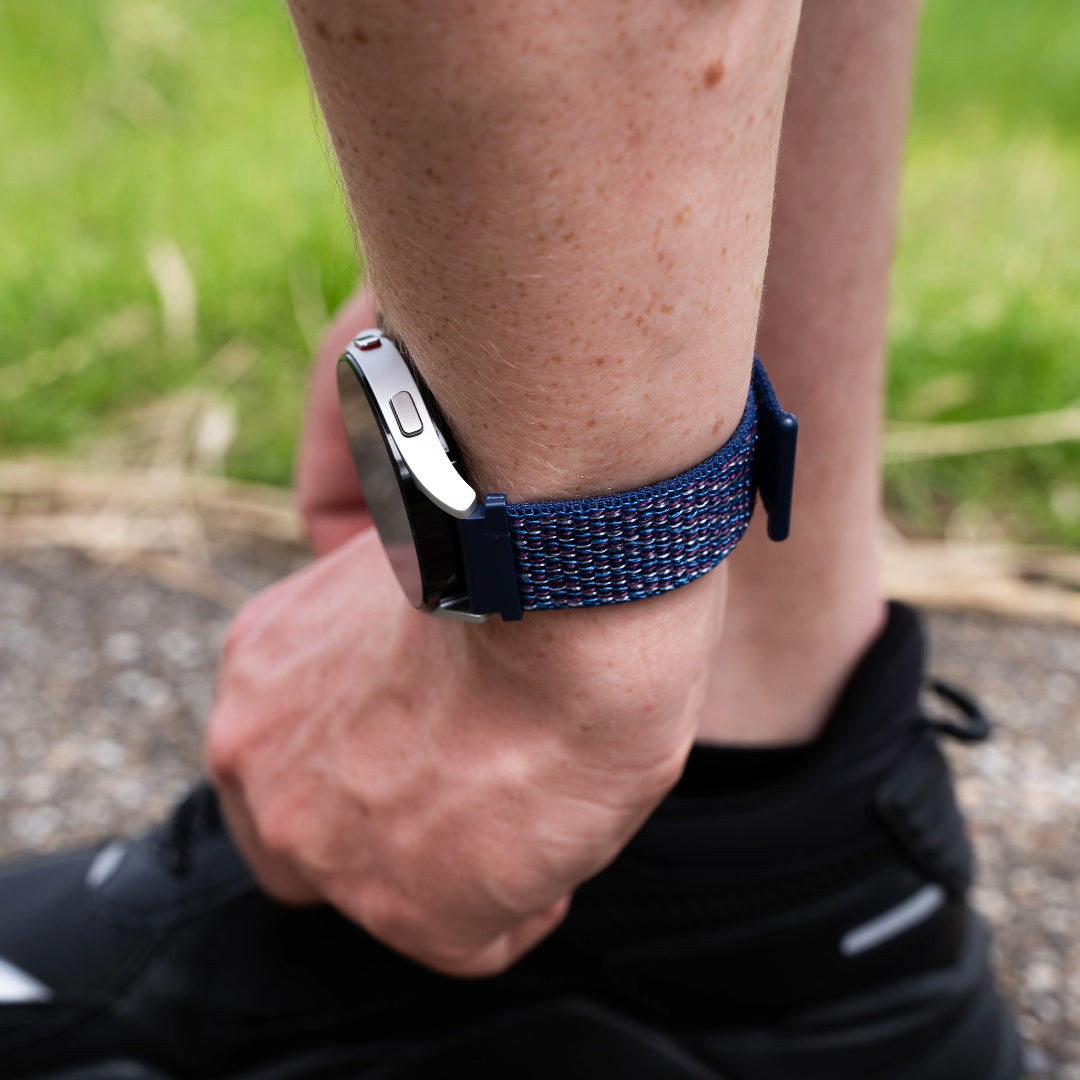Person, die eine Smartwatch mit blauem gewebtem Armband trägt. Die Person trägt einen schwarzen Schuh. Der Hintergrund ist verschwommen grün.