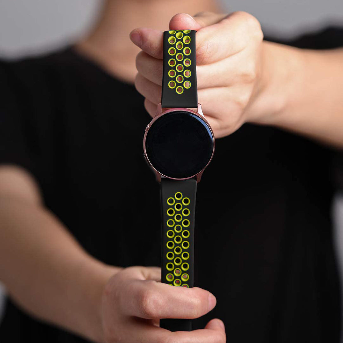 Eine Person hält eine Smartwatch. Die Uhr hat einen schwarzen Bildschirm und ein schwarzes Armband mit gelben Kreisen. Der Hintergrund ist unscharf.