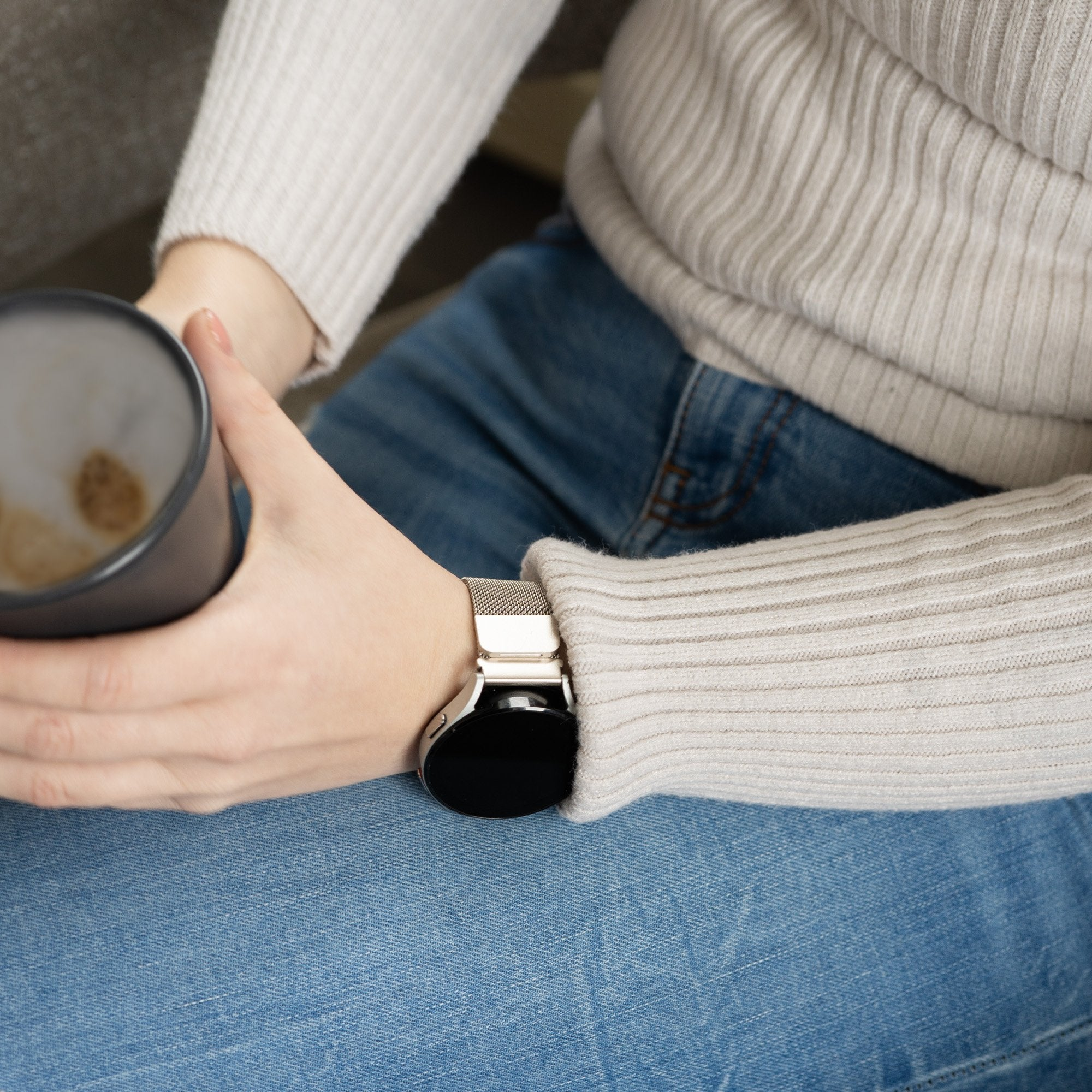 Person hält Kaffeetasse, trägt eine Uhr und Jeans. Der Pullover ist beige.