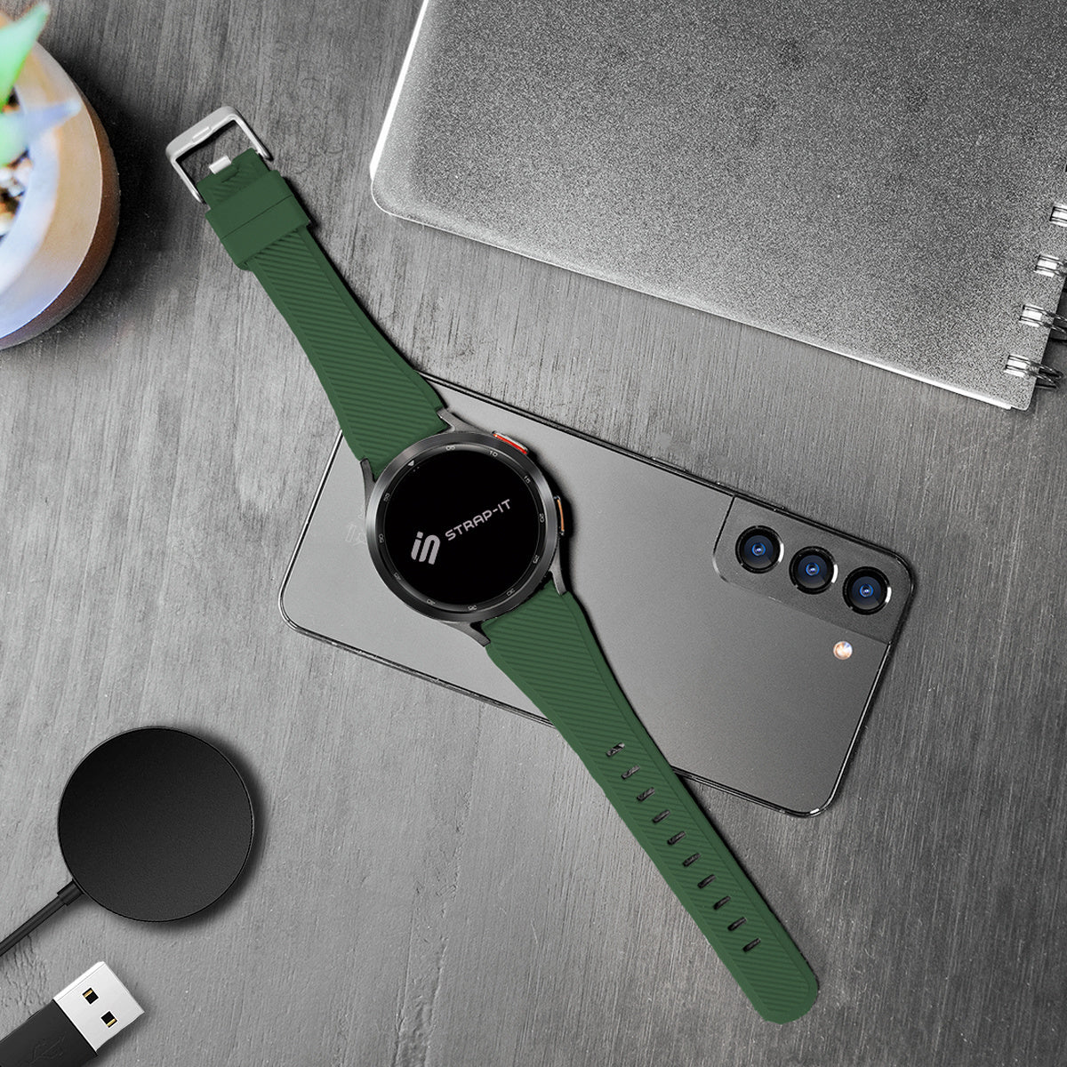 Eine grüne Smartwatch mit grünem Armband liegt auf einem grauen Telefon. Ein kabelloses Ladegerät und ein Notizbuch sind ebenfalls vorhanden.