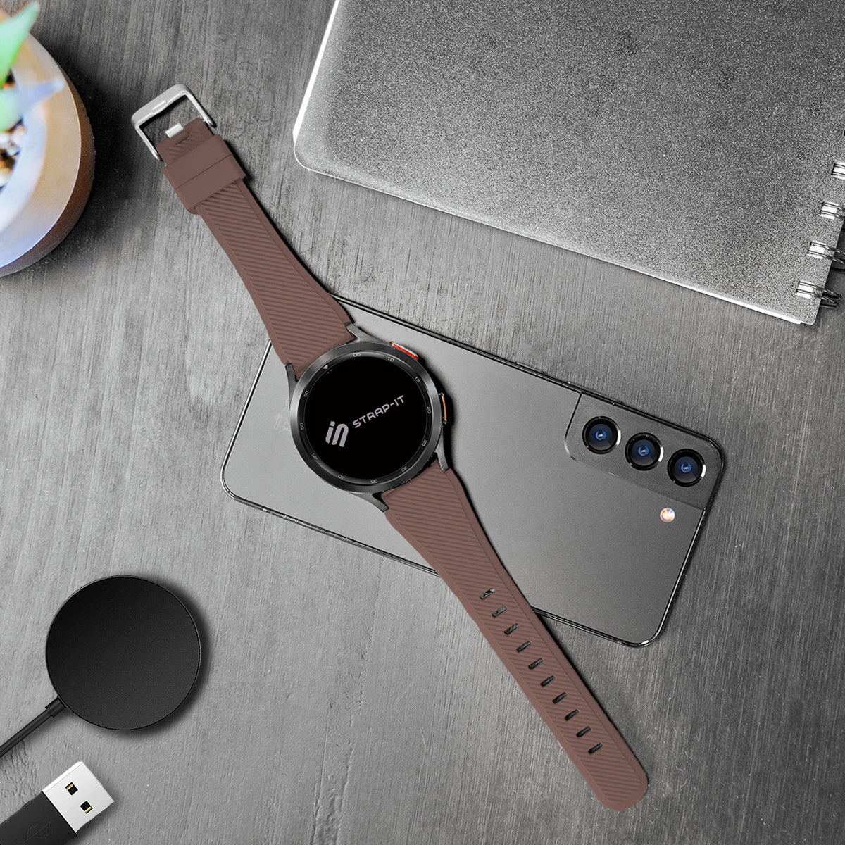 Eine braune Smartwatch mit passendem Armband liegt auf einem grauen Telefon. Ein Ladegerät und ein Notizbuch befinden sich ebenfalls auf einer Holzoberfläche.