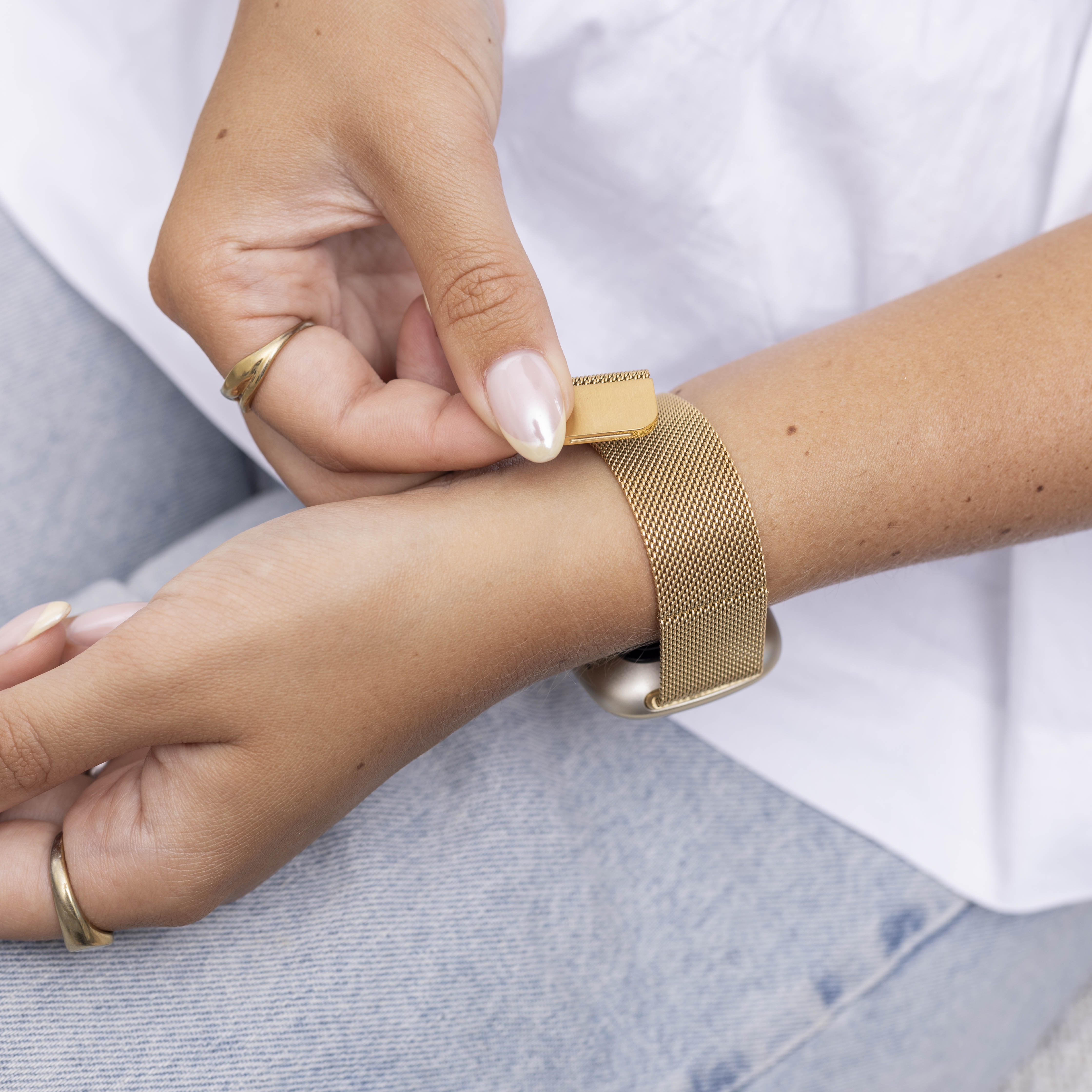 Eine Hand passt ein goldenes Armband an. Die Person trägt einen Ring und hat gepflegte Nägel. Der Hintergrund zeigt ein weißes Hemd und Jeans.