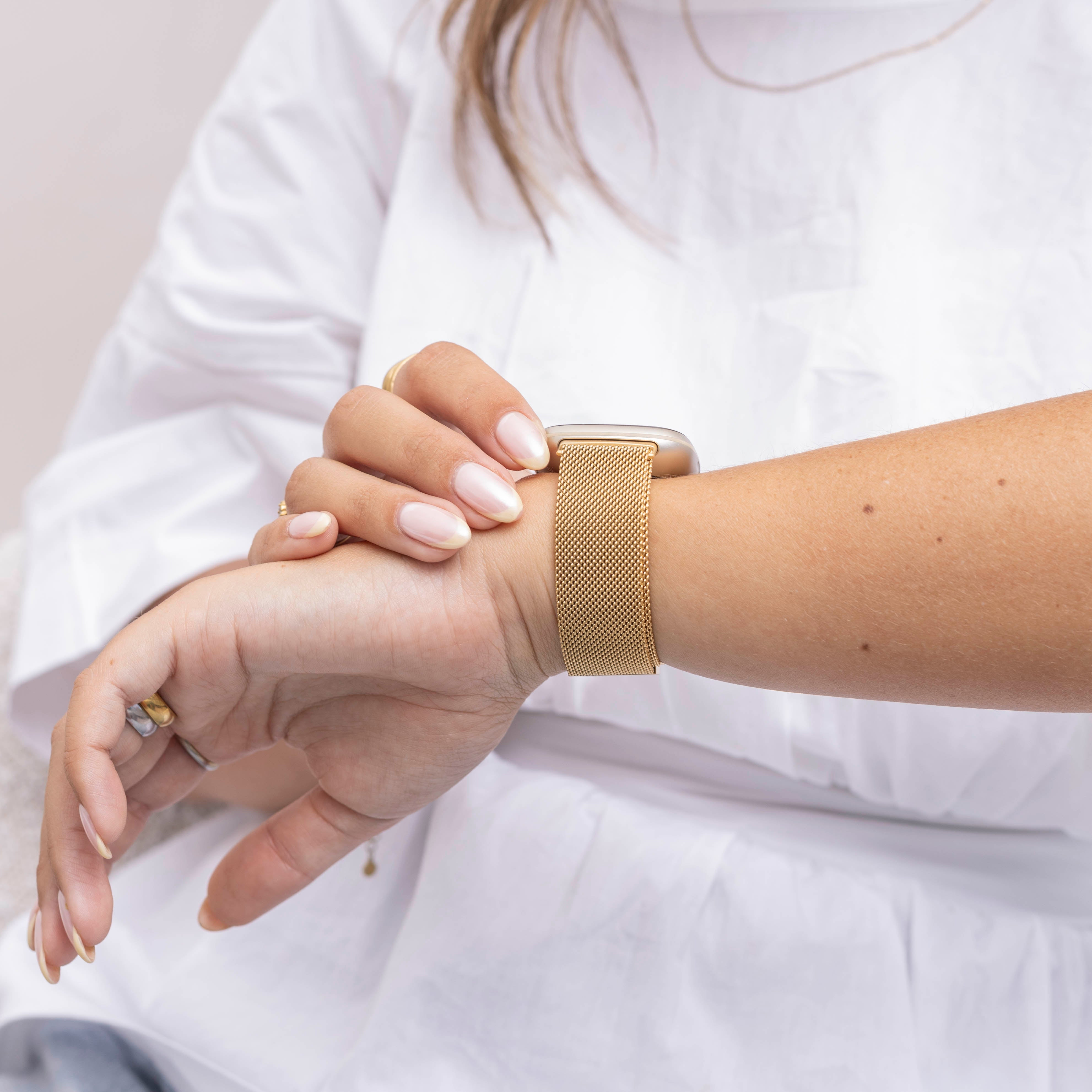 Hand einer Person mit langen Nägeln, die eine goldene Uhr trägt. Die Person trägt ein weißes Hemd.