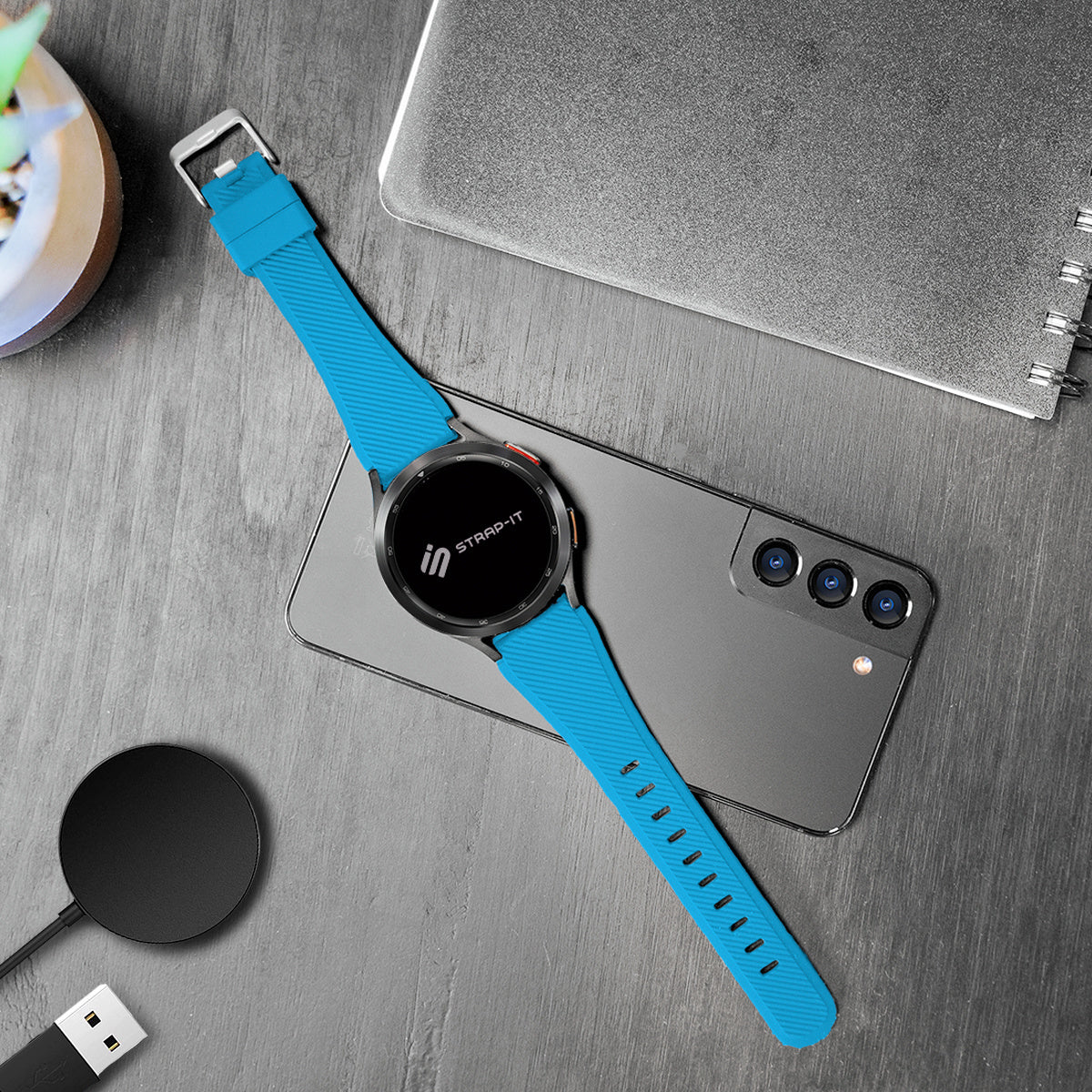 Eine Smartwatch mit blauem Armband liegt auf einem Telefon. Weitere Artikel sind ein kabelloses Ladegerät, ein Notizbuch und eine Pflanze.