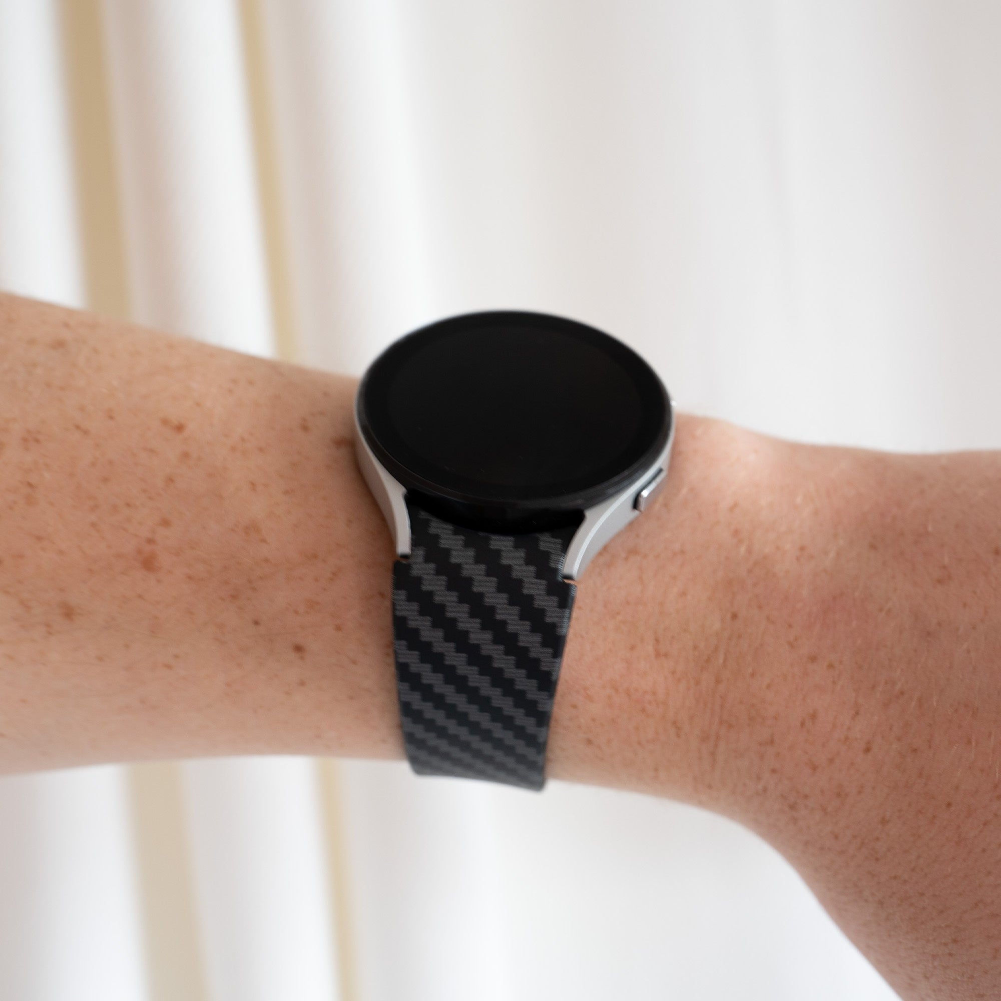 Ein Arm mit einer schwarzen Smartwatch. Die Uhr hat ein silbernes Gehäuse und ein gemustertes schwarzes Armband. Hintergrund ist ein weißer Vorhang.