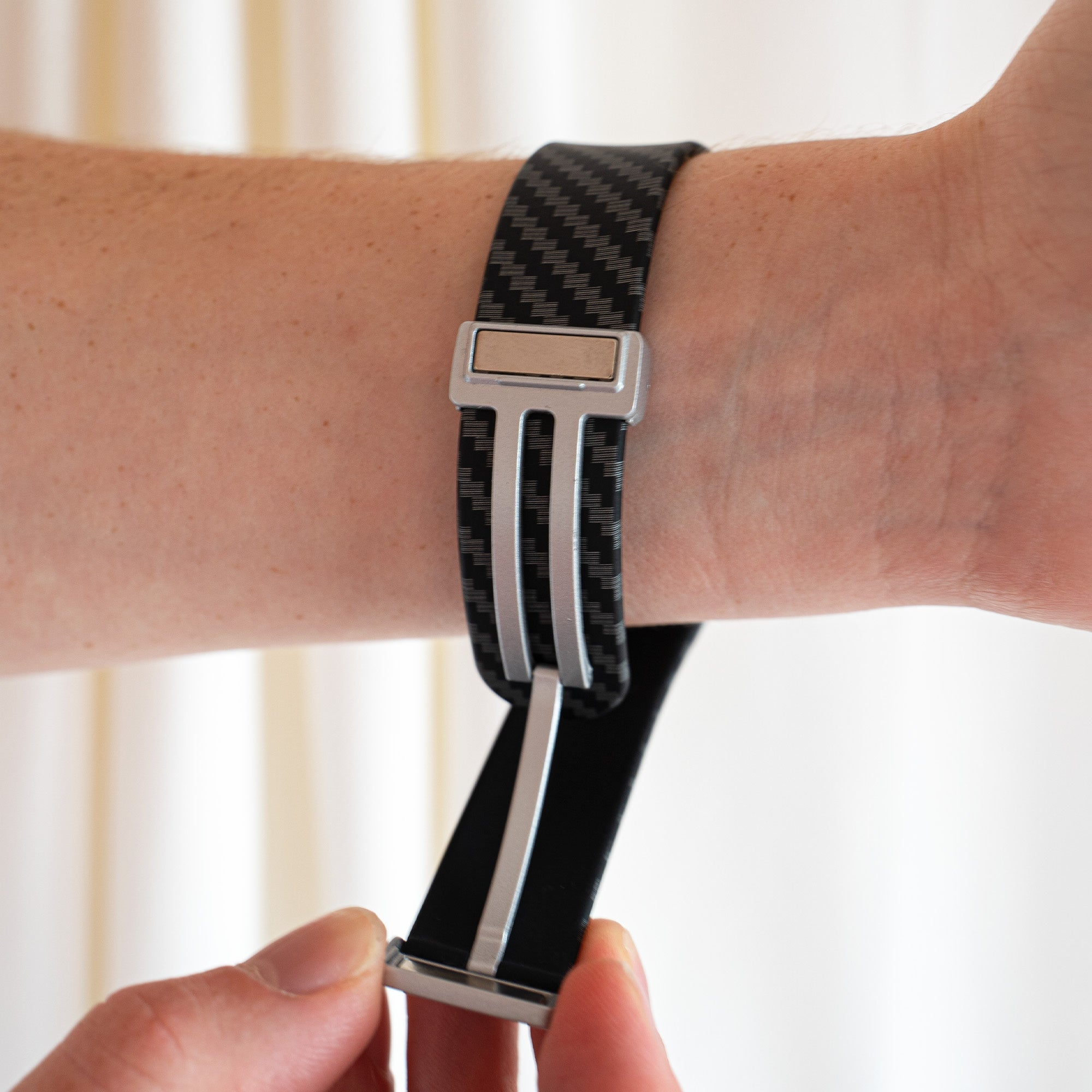 Ein Handgelenk mit schwarzem Uhrenarmband. Das Band hat ein Karbonfasermuster und einen Metallverschluss. Eine Hand hält das Band.