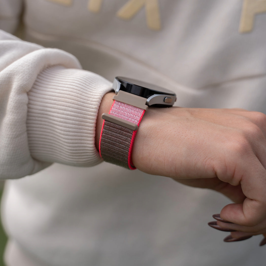 Ein Handgelenk mit einer Smartwatch. Die Uhr hat ein rosa-braunes Armband. Der Hintergrund ist verschwommen, ein weißer Pullover ist sichtbar.