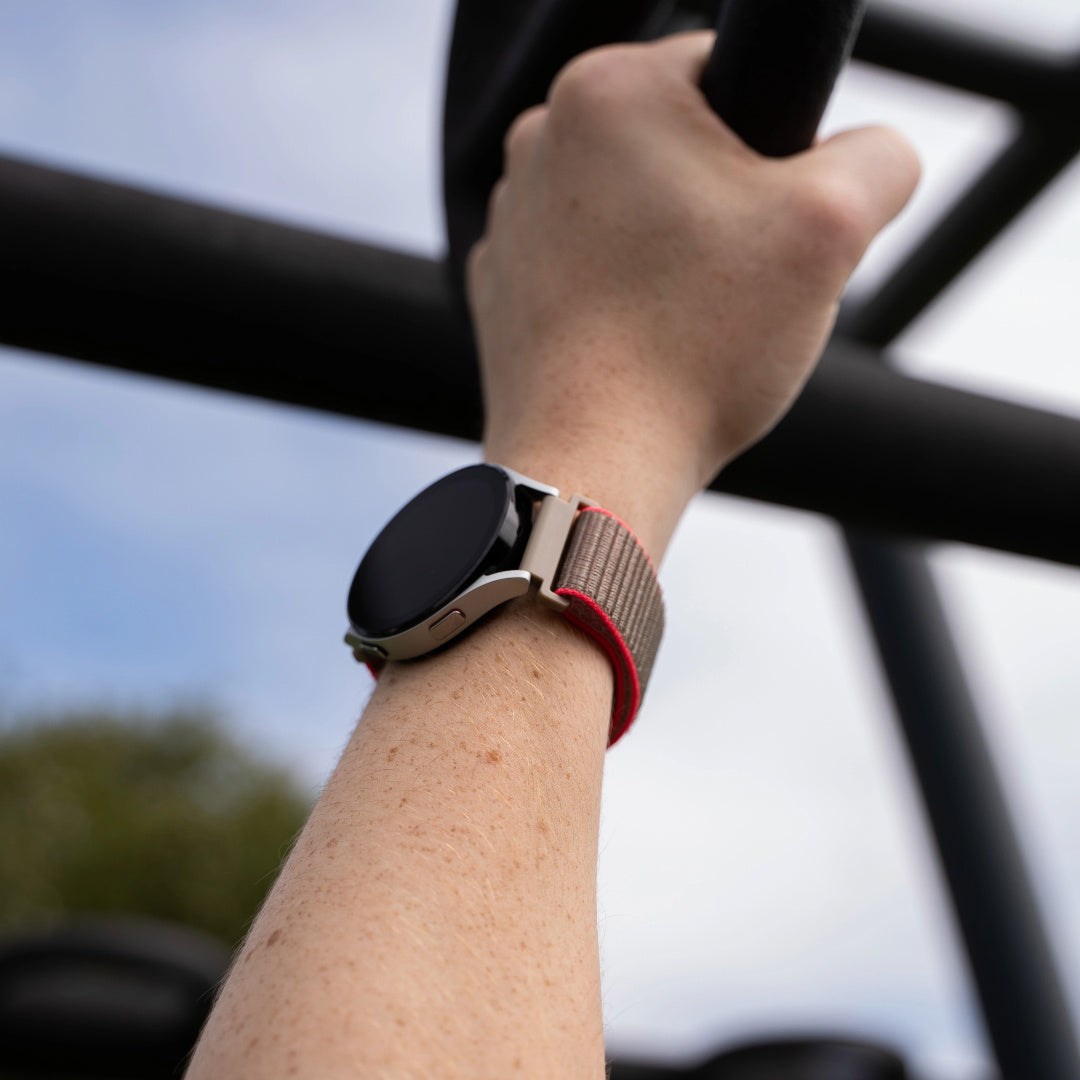 Arm einer Person, die eine schwarze Stange hält, eine Smartwatch am Handgelenk. Die Uhr hat ein graues Gehäuse und ein braunes, rot gefüttertes Armband.